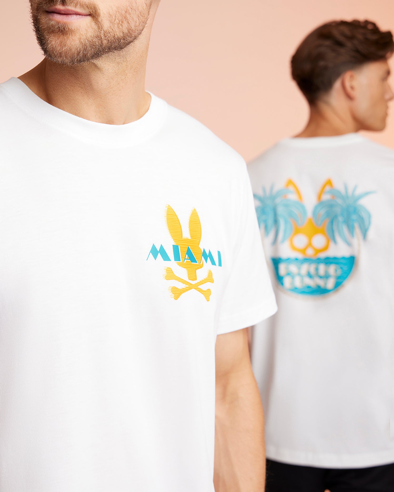 Two men wearing white men's Florida tees with colorful graphics. The man in the foreground has a Psycho Bunny MENS MIAMI TEE - B6U550W1PC that says 