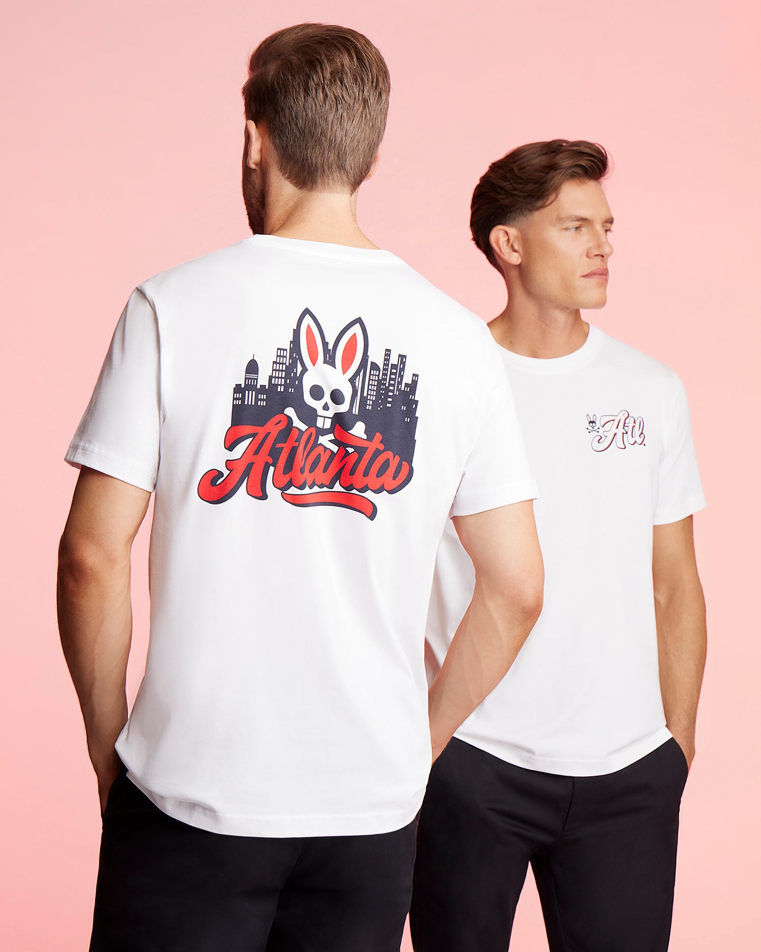 Two men are wearing matching Psycho Bunny MENS ATLANTA CITY TEE - B6U966X1PC tees in luxurious Pima cotton jersey. The back of the shirts features a cartoon skull with bunny ears, a cityscape, and the word 