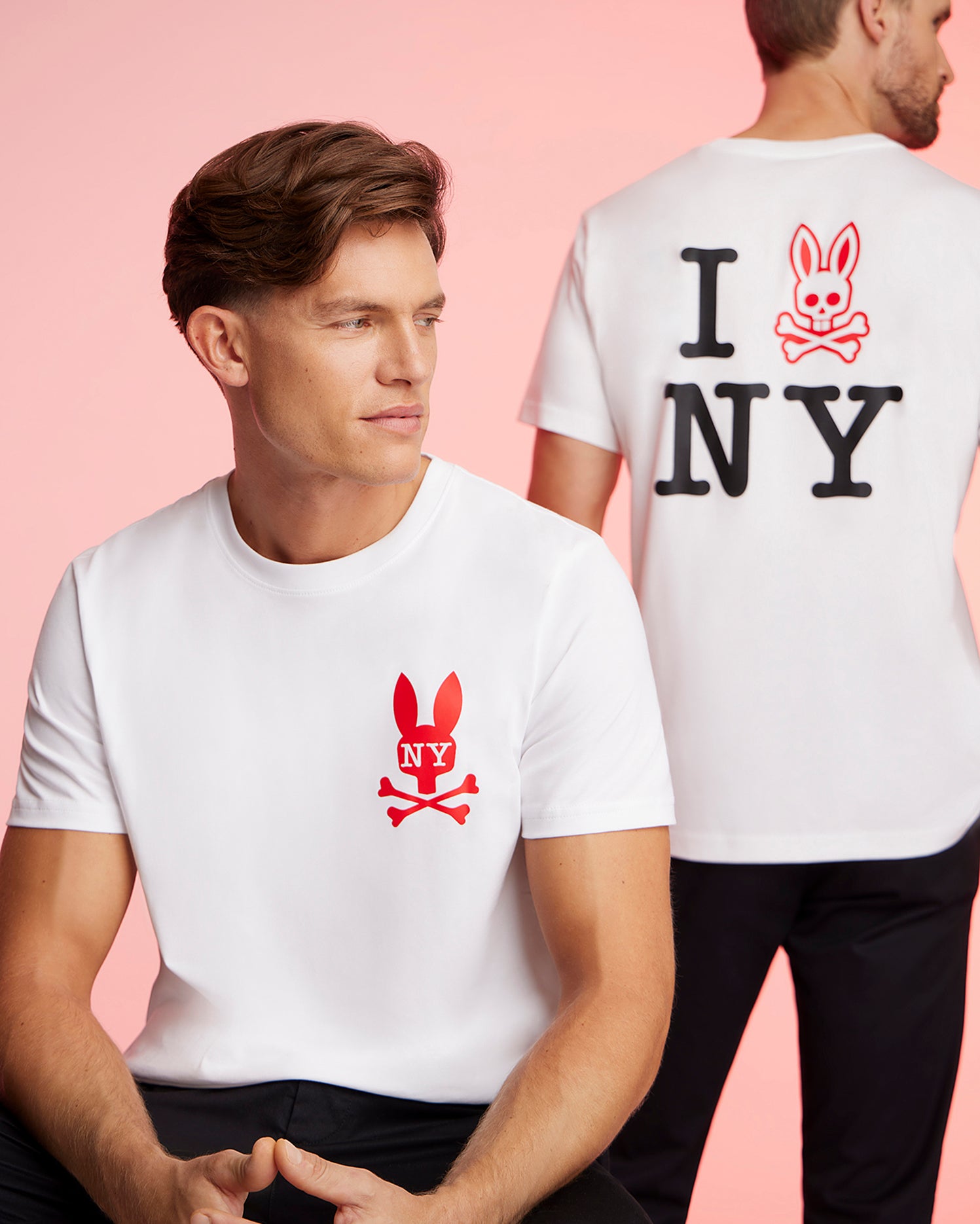 Two men wearing matching Pima cotton jersey white t-shirts with a red bunny and crossbones logo. The man in the foreground faces forward, while the man in the background faces away, revealing text on his Psycho Bunny MENS NEW YORK TEE - B6U554W1PC that reads 