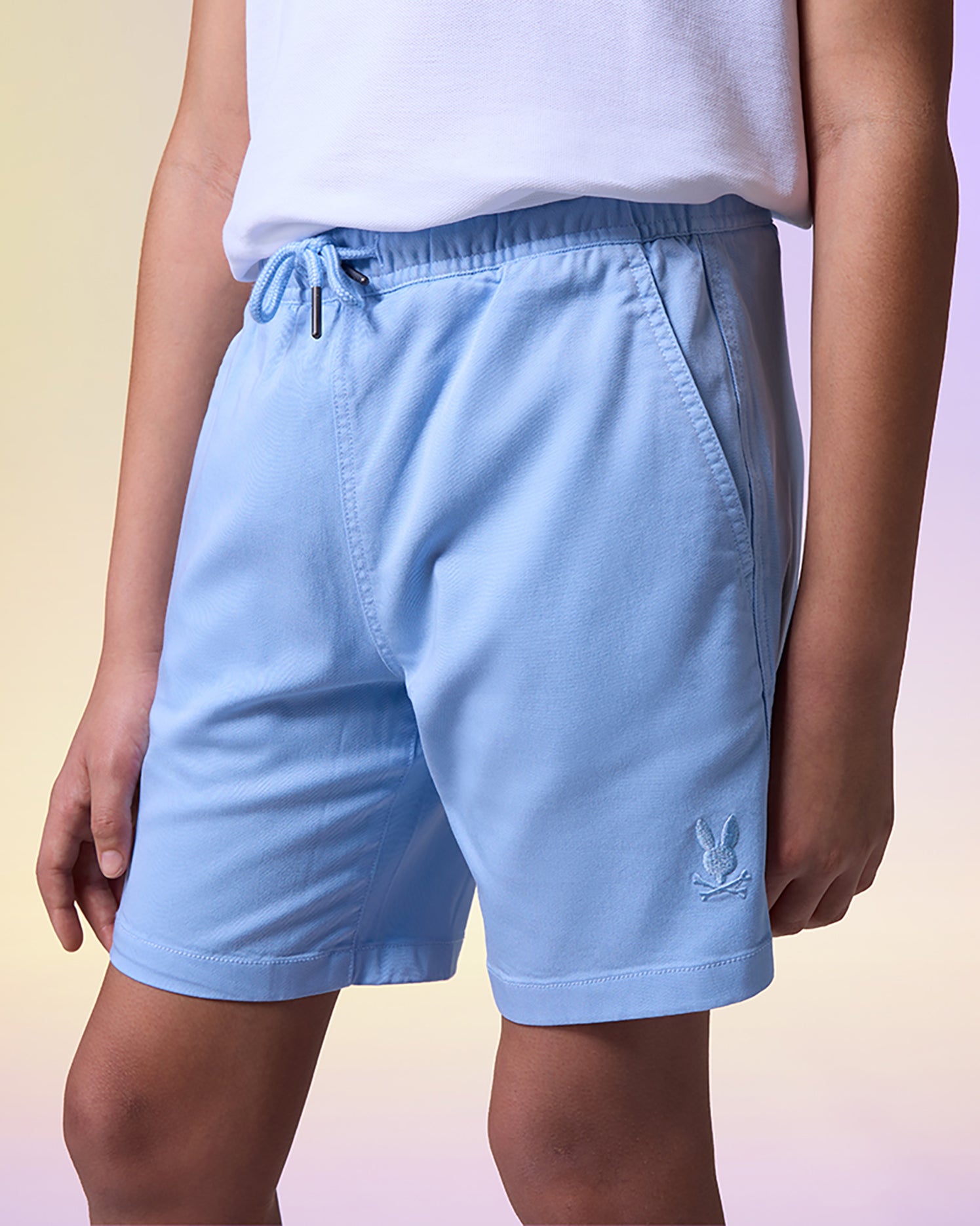 A person stands wearing a white shirt and light blue KIDS WILLIS STRETCH TENCEL SHORT - B0R239Y1WB by Psycho Bunny with pockets and an embroidered logo on the left leg, made from super-soft stretch Tencel-blend twill. The background is a gradient of soft pastel colors.