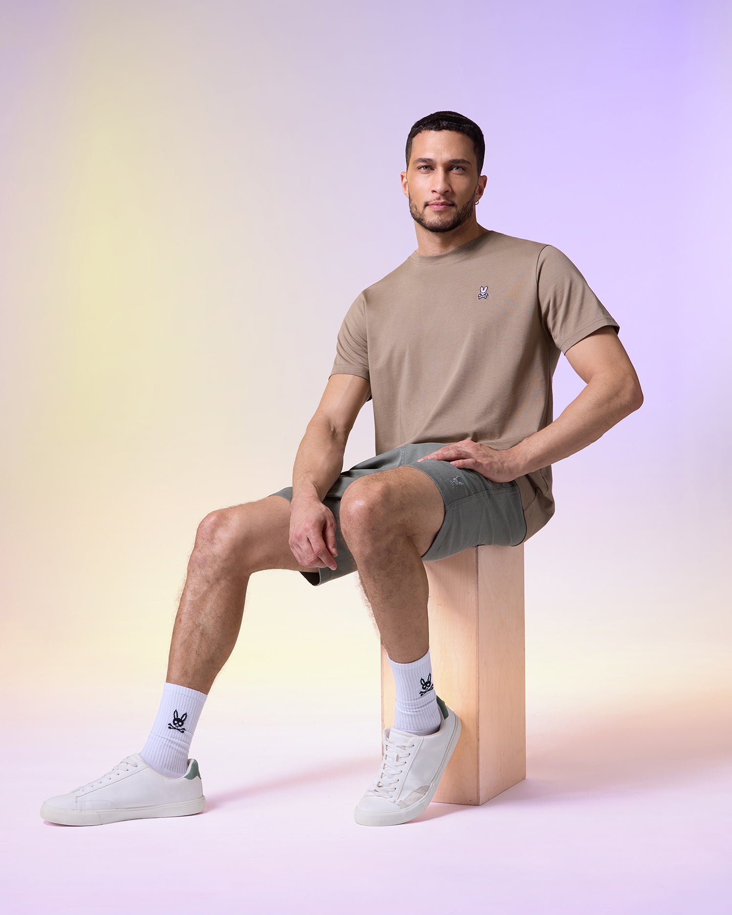 A man sits casually on a wooden stool, wearing a Psycho Bunny Men's Classic Crew Neck Tee in beige, gray shorts, and white sneakers, with a soft colorful gradient background.