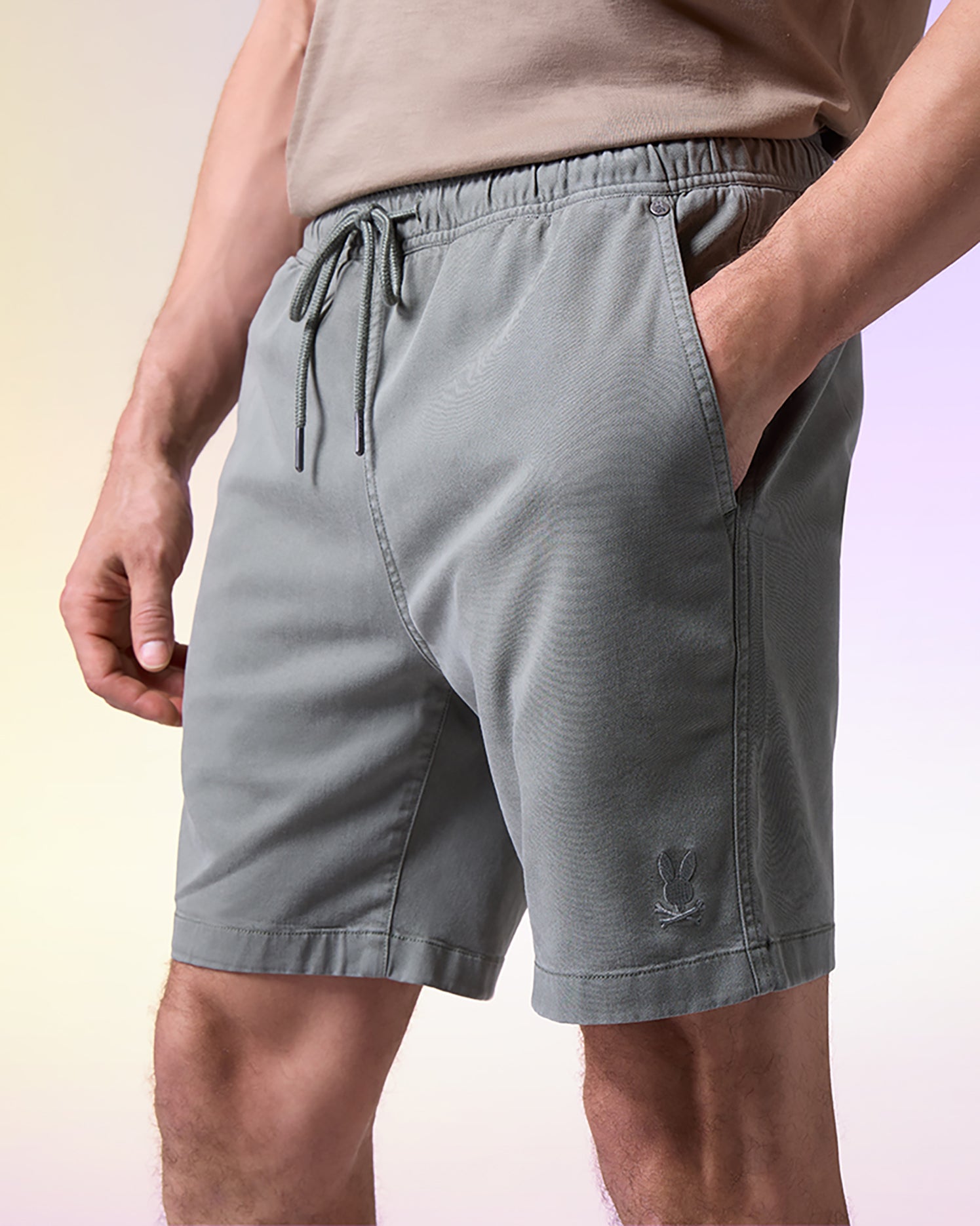 A close-up of a man wearing gray Psycho Bunny MENS WILLIS STRETCH TENCEL SHORT - B6R239Y1WB drawstring shorts with a small logo on the left thigh, standing against a soft gradient background.