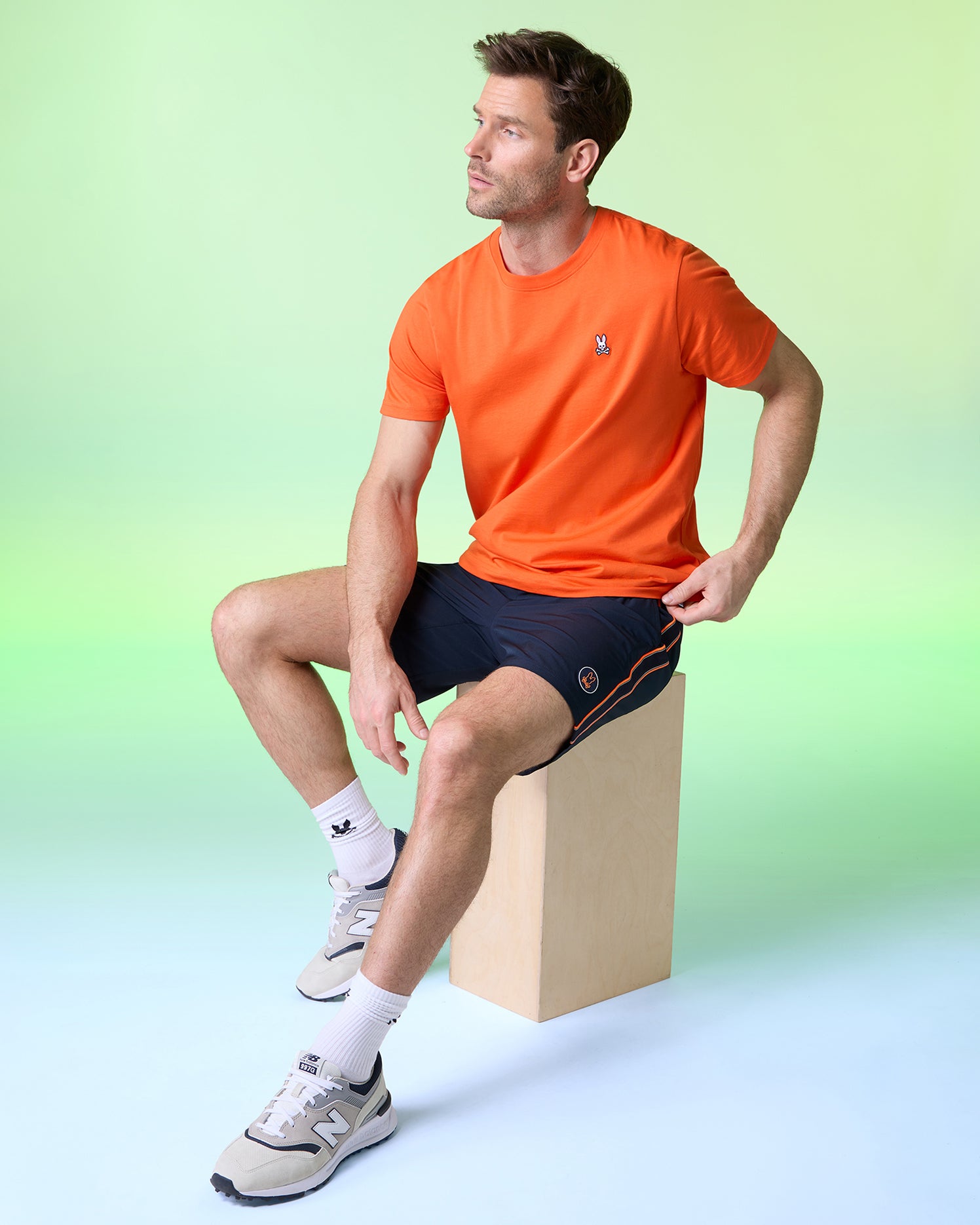 A man sits on a wooden block against a gradient green background. He wears an orange Psycho Bunny men's MENS CLASSIC CREW NECK TEE - B6U014CRPC with an embroidered Bunny logo, navy shorts, white socks, and gray sneakers. His hands rest on his thighs as he gazes to the side.