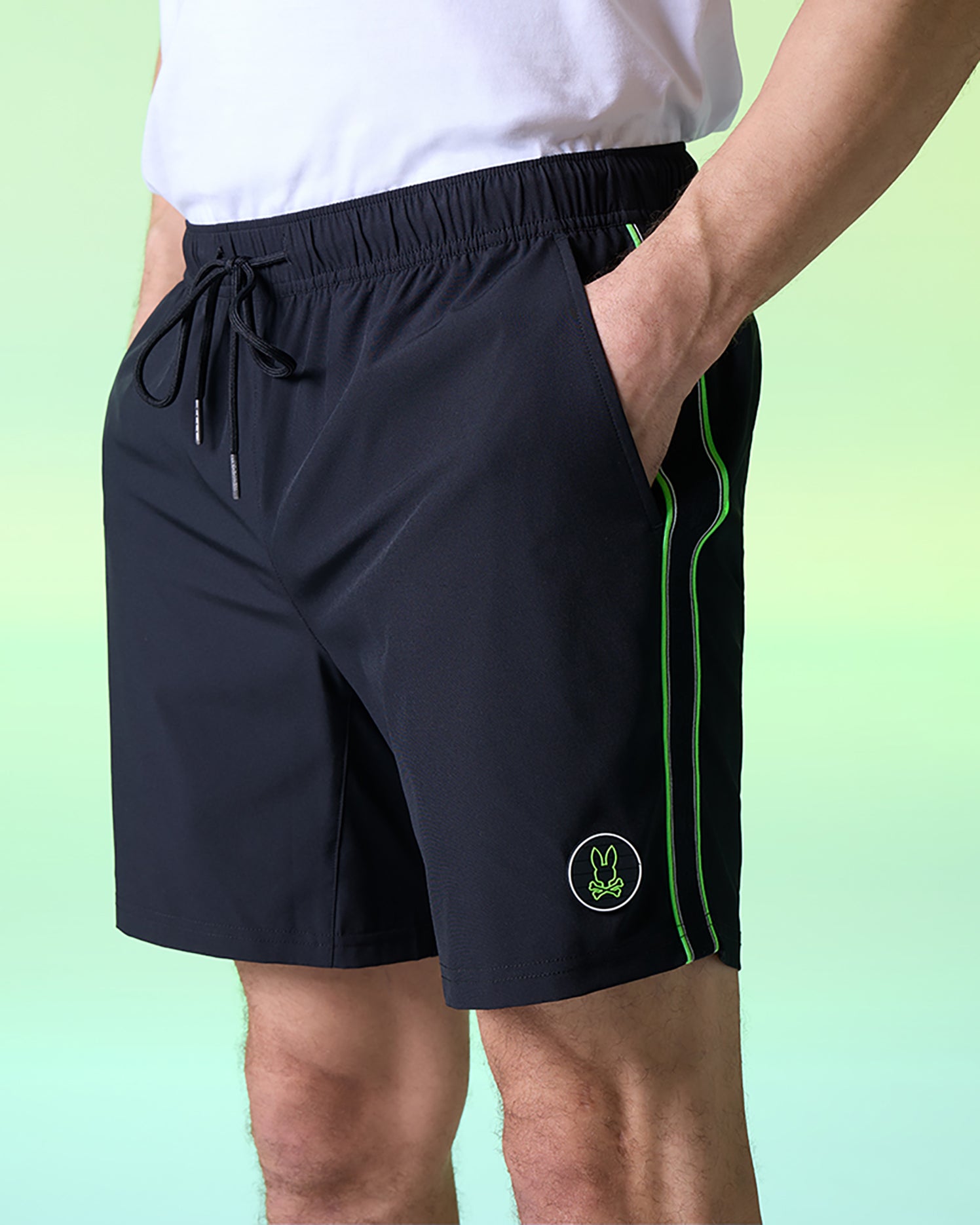 A person is standing wearing black Psycho Bunny MENS SONTERRA STRIPE SPORT SHORT - B6R464C200 made from breathable fabric, featuring green piping along the sides and a small green bunny logo on the left thigh. The background is a gradient of light green and light blue. The person is also wearing a plain white shirt.
