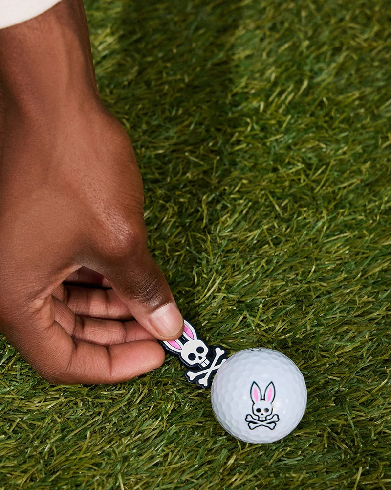 A hand places a Psycho Bunny BALL MARKER - B6A698C200, adorned with a white bunny skull and crossbones, on grass next to a matching bunny skull and crossbones golf tee from the golfers collection.