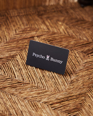 A stylish black Digital Gift Card from Psycho Bunny, featuring a pink bunny skull logo, lies gracefully on a herringbone-patterned woven surface. Ideal for gifting, it can be effortlessly redeemed at checkout for your preferred styles.