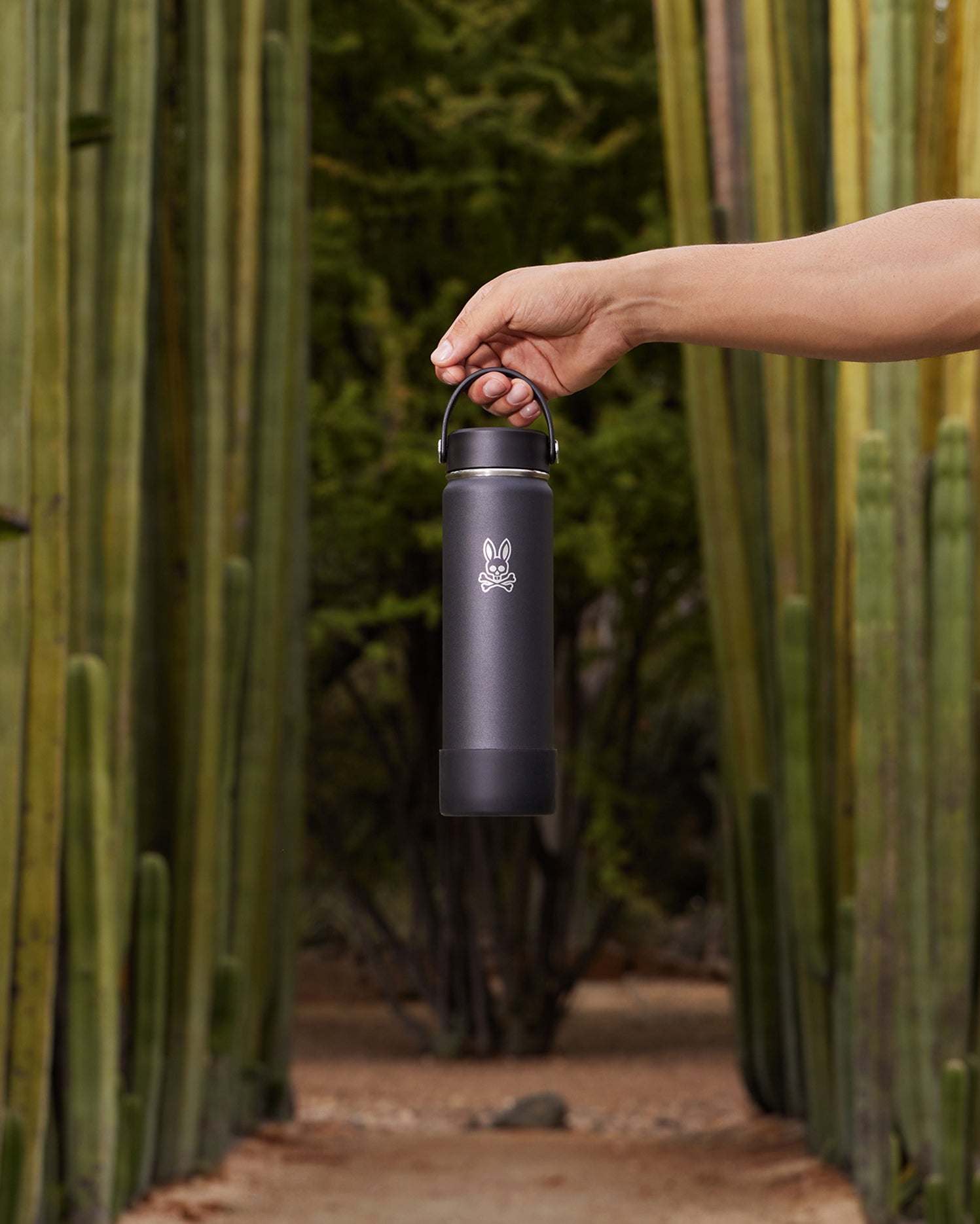 A person's hand holding a Psycho Bunny insulated stainless steel water bottle (B6A909U1BO) by its handle in front of tall cactus plants in a desert setting. The bottle features a logo of a rabbit.