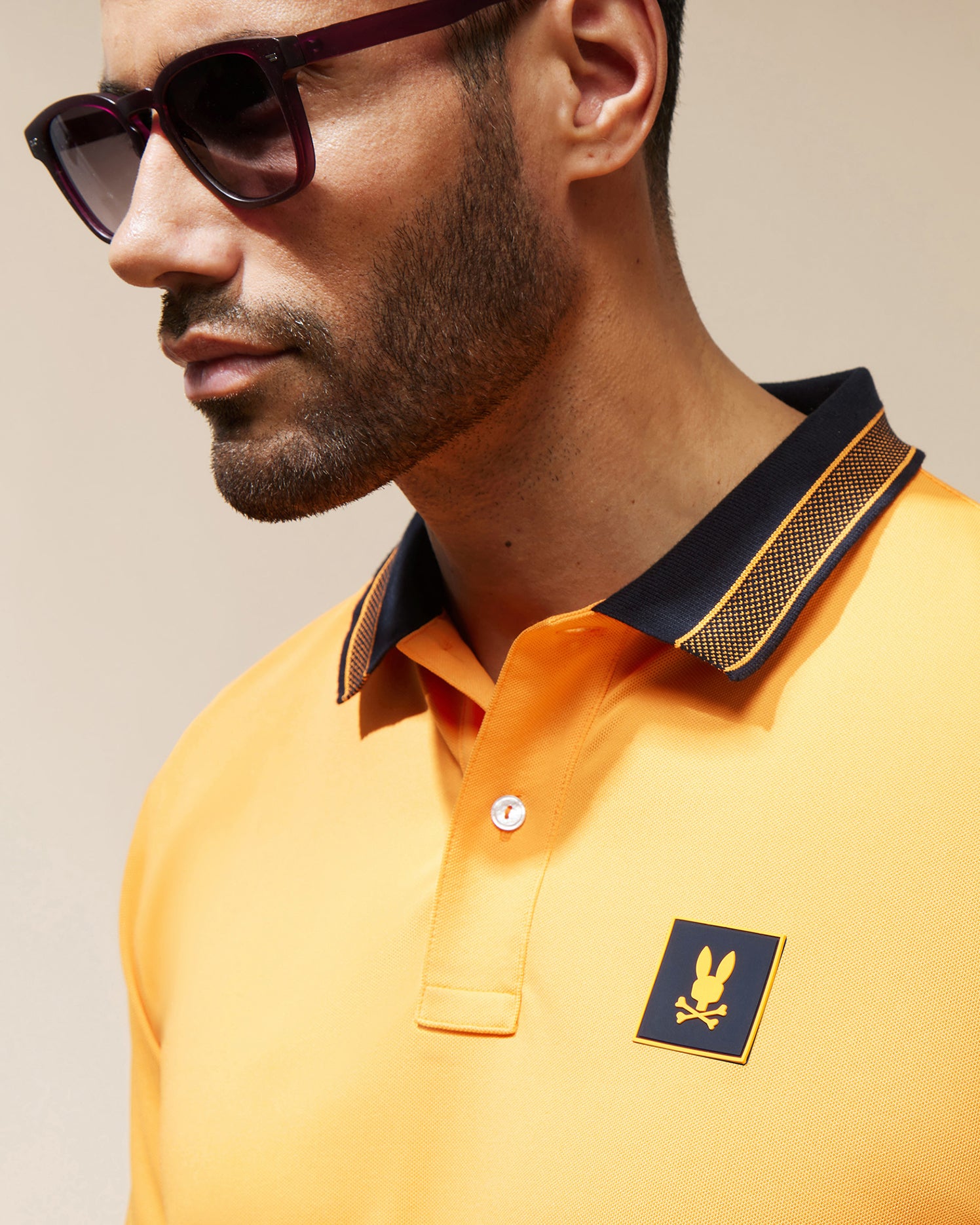 A man with a dark beard, wearing sport-forward sunglasses and an orange MENS TARRYTOWN SPORT POLO - B6K333B200 from Psycho Bunny, featuring a black and yellow collar, gazes to the side. The polo, made of breathable material, showcases a bunny head with crossbones on the chest. The background is a neutral tone.