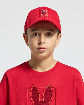 A young person stylishly sporting the KIDS PIERCE BASEBALL CAP - B0A183E200 by Psycho Bunny paired with a red T-shirt featuring a rabbit graphic. The cotton comfort ensemble is set against a plain light gray background.