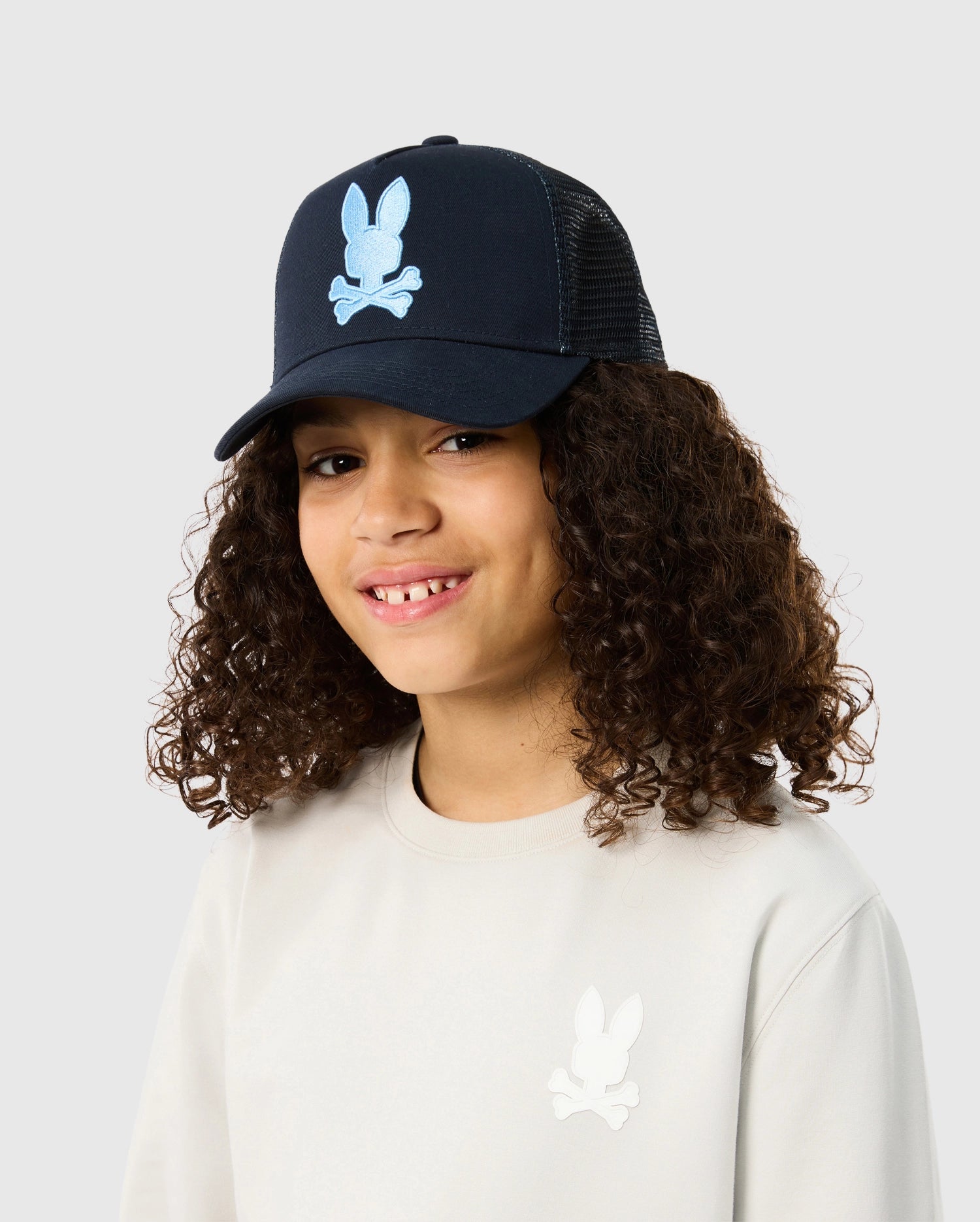 A young person with curly hair is smiling and wearing a beige sweatshirt with a bunny logo. They are also wearing a dark blue KIDS HOUSTON TRUCKER HAT - B0A550C2HT by Psycho Bunny featuring the same bunny logo in light blue, complete with snapback fastening for the perfect fit. The background is plain and light-colored.