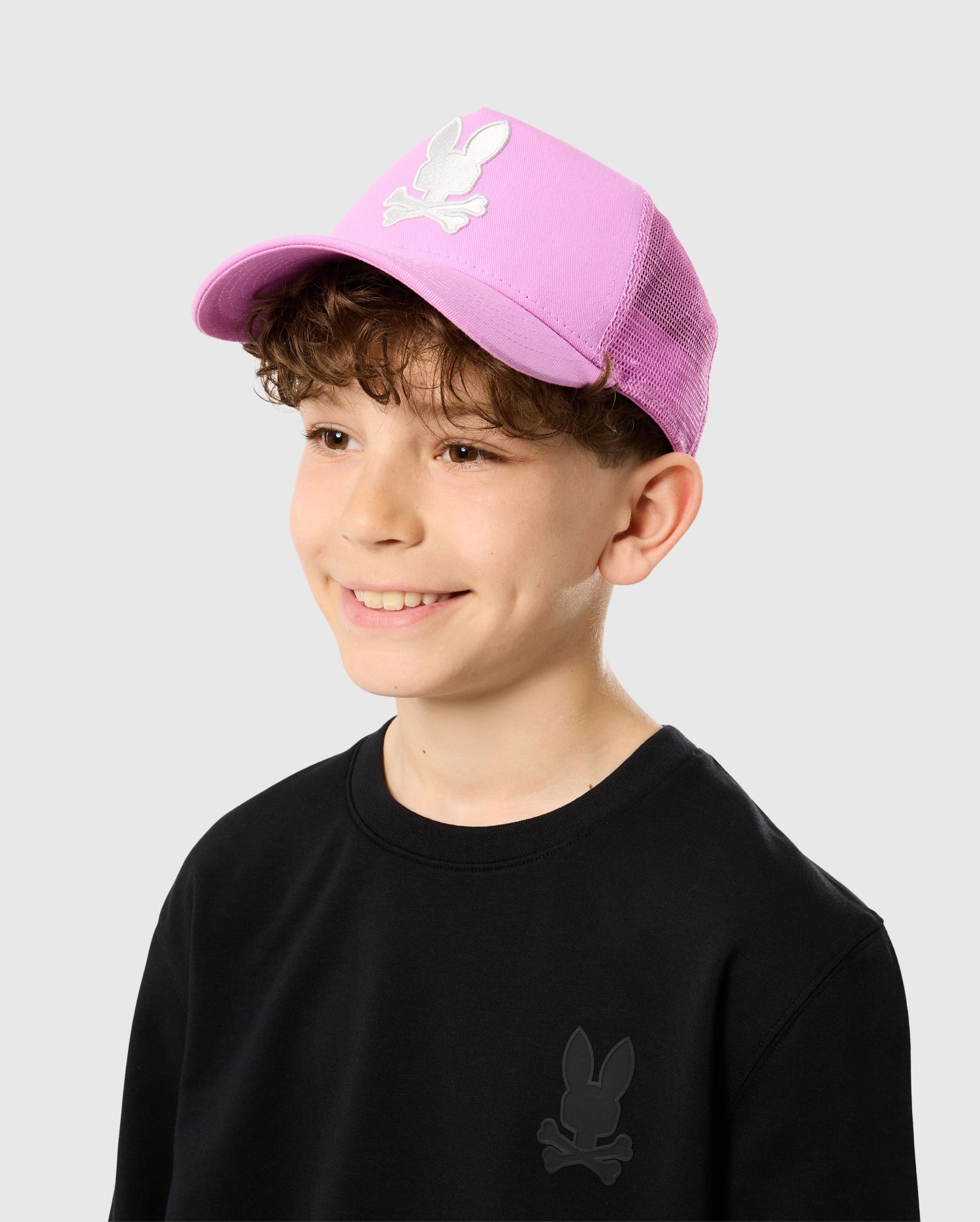 A young boy with curly hair is smiling and looking to the side. He is wearing a black sweatshirt with a small bunny logo on it and a pink Psycho Bunny KIDS HOUSTON TRUCKER HAT - B0A550C2HT featuring a similar white bunny logo with crossed bones underneath. The background is plain light grey.
