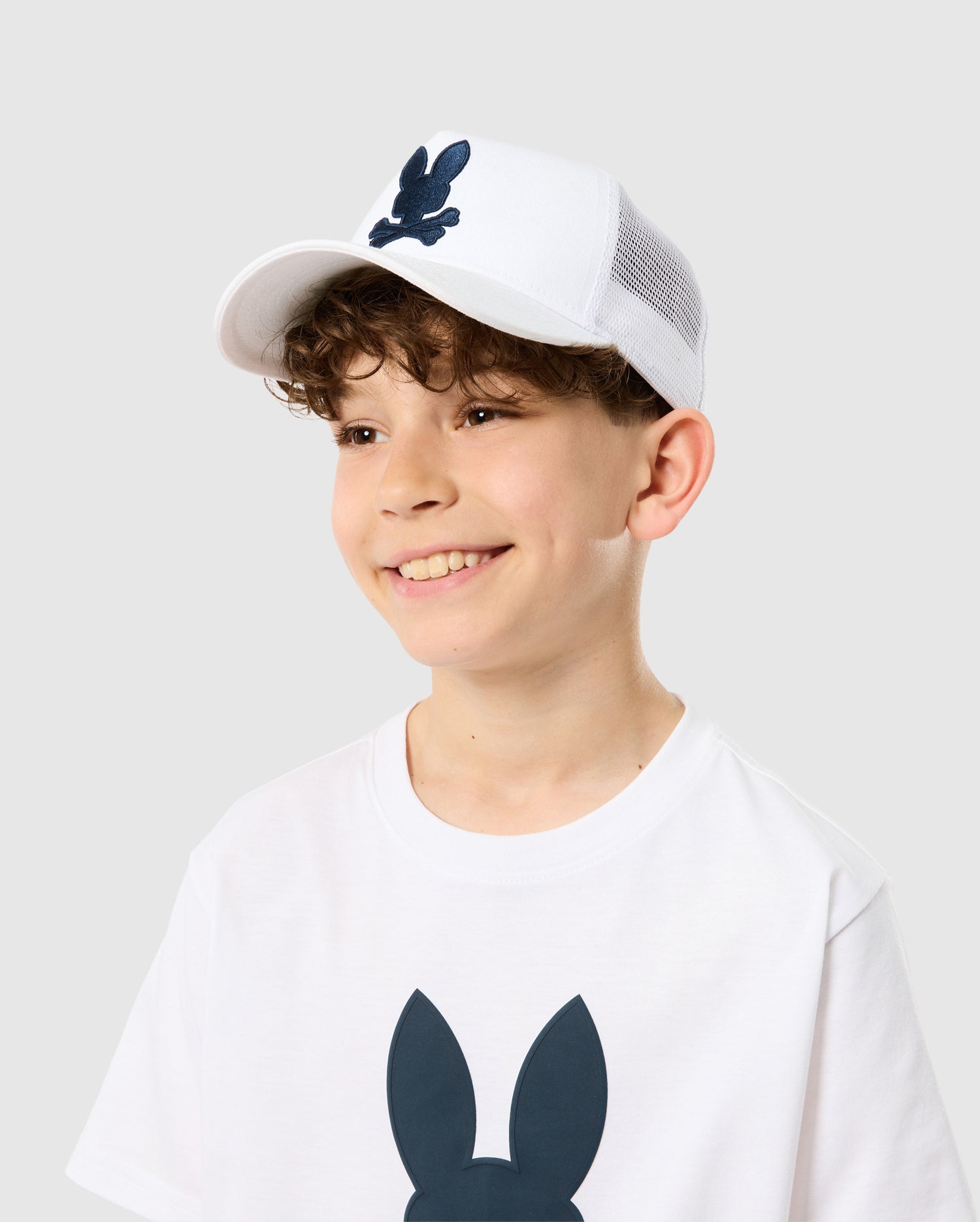 A young boy wearing a white Psycho Bunny Houston trucker cap with snapback fastening and t-shirt with a blue bunny design, smiling against a plain background. His curly hair peeks out from under the cap.