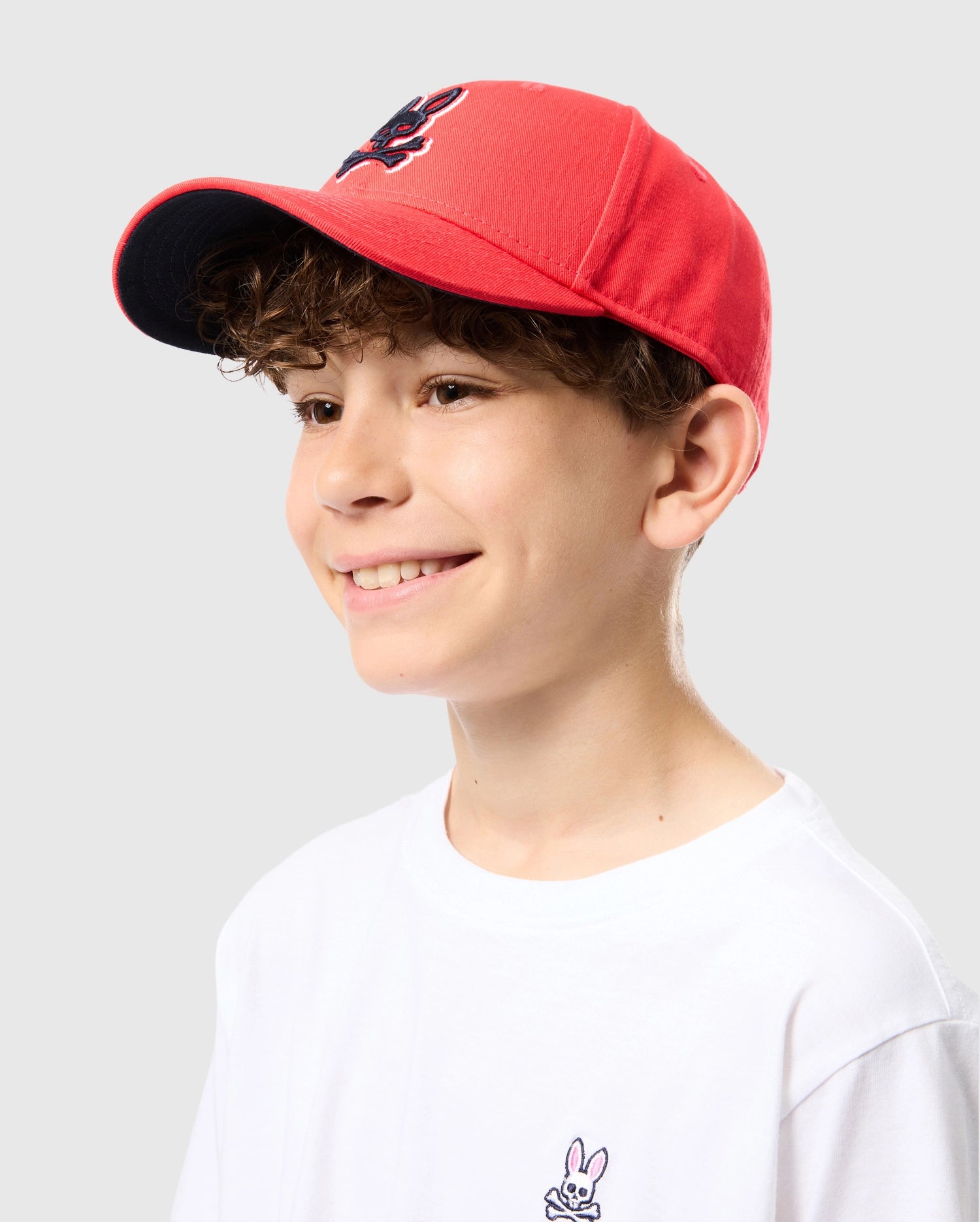 A smiling young person with curly hair wears a red KIDS KAYDEN BASEBALL CAP - B0A677C200 from Psycho Bunny, featuring a black brim and a navy blue logo on the front. They are also wearing a white T-shirt with a small bunny logo on the chest, set against a plain light gray background.