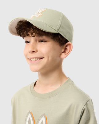 A smiling young boy with curly brown hair is wearing a Psycho Bunny KIDS KAYDEN BASEBALL CAP - B0A677C200 in light green, featuring an embroidered Bunny logo, paired with a matching light green t-shirt. The background is plain and light-colored.