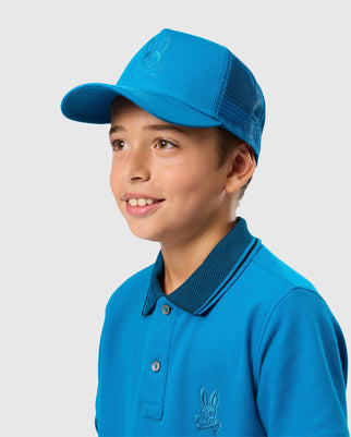 A smiling young boy is wearing the KIDS MALTON TRUCKER CAP by Psycho Bunny, featuring bright blue fabric and mesh ventilation, along with a matching cotton polo shirt that showcases a small embroidered logo on the chest. He is looking slightly to the side against a plain, light-colored background.