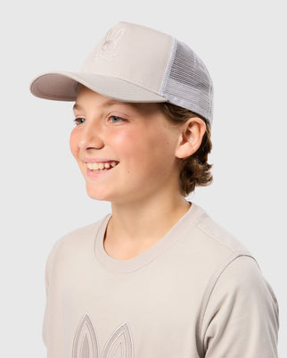 A young person wearing a Psycho Bunny KIDS MALTON TRUCKER CAP (B0A797D200) in beige, featuring mesh ventilation and a subtle embroidered design on the front. They are also wearing a matching beige cotton t-shirt with a similar embroidered design. The plain gray background highlights their smile as they look to the side.