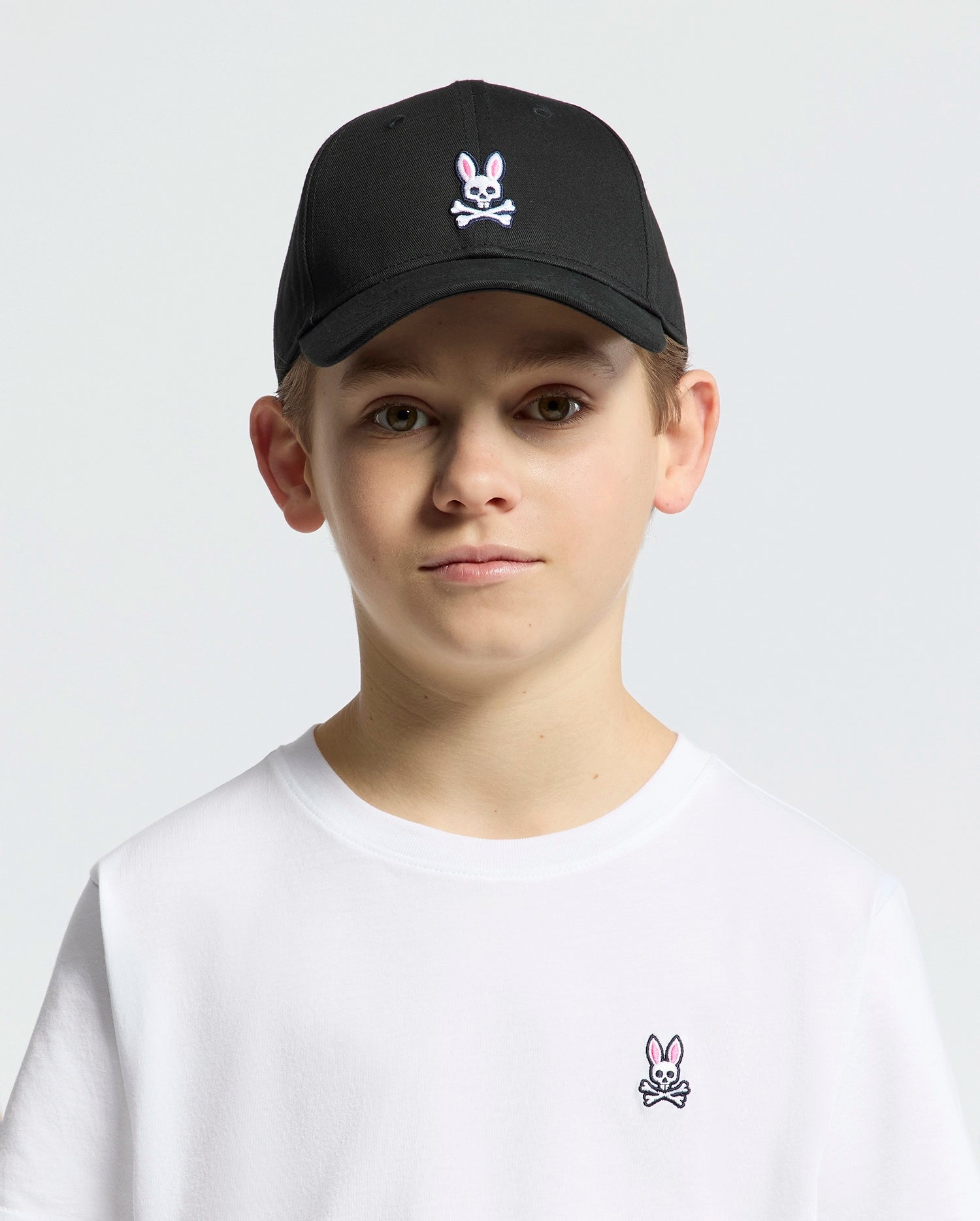 A young boy wears a KIDS CLASSIC BASEBALL CAP - B0A816A1HT by Psycho Bunny, showcasing an adjustable fit with a pink and white embroidered bunny skull. His matching white T-shirt also displays the emblem against a plain white background.