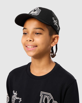 A person with braided hair is smiling, wearing the sporty black KIDS ZACK BASEBALL CAP - B0A886D200 by Psycho Bunny, crafted from 100% cotton, along with a matching black shirt. The background is plain light gray.