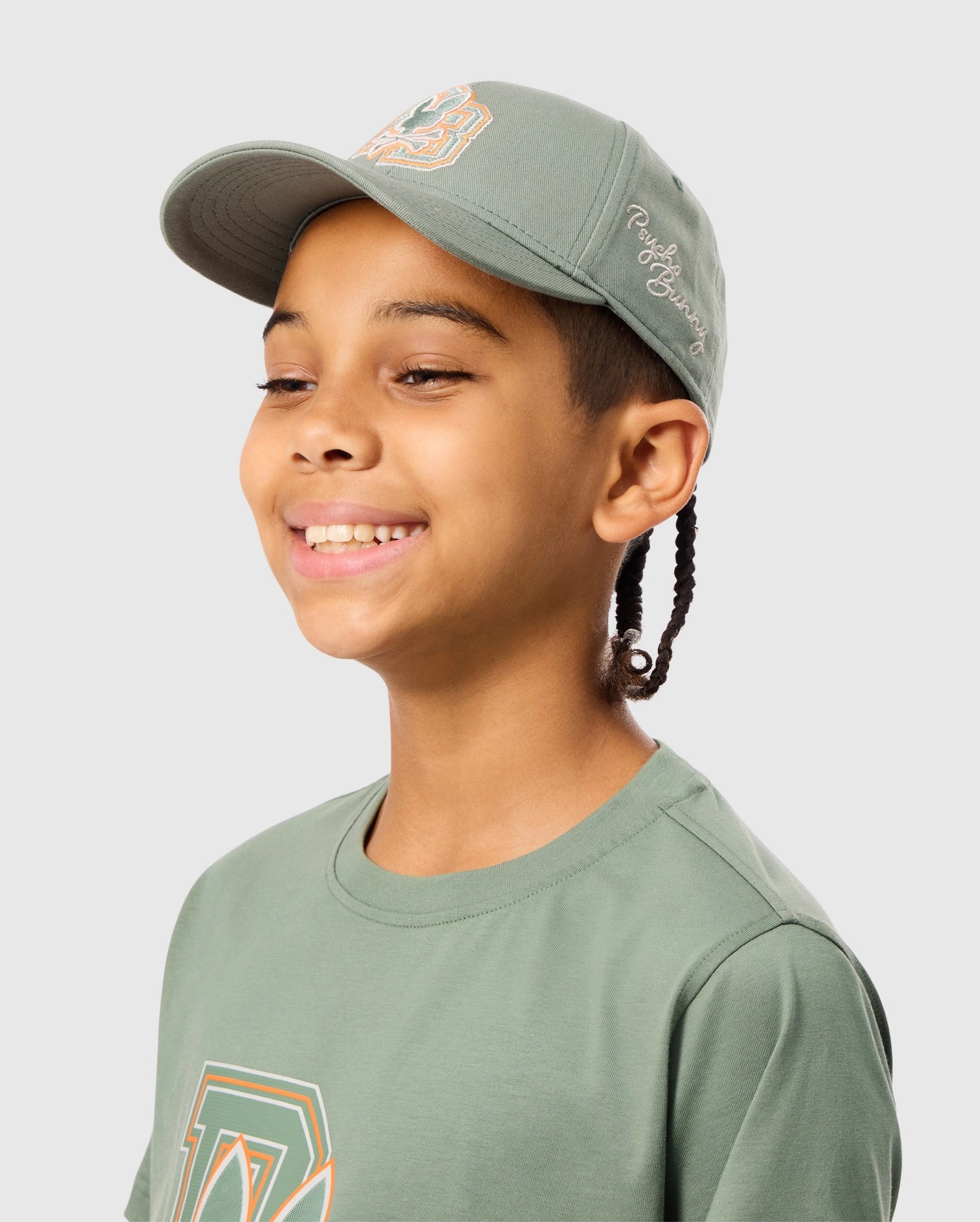 A sporty child beams in a green KIDS ZACK BASEBALL CAP from Psycho Bunny, paired with a matching T-shirt, both proudly displaying logos. The woven cotton cap features embroidered designs on the front and side. The child, with braids, gazes slightly to the side against a light gray background.