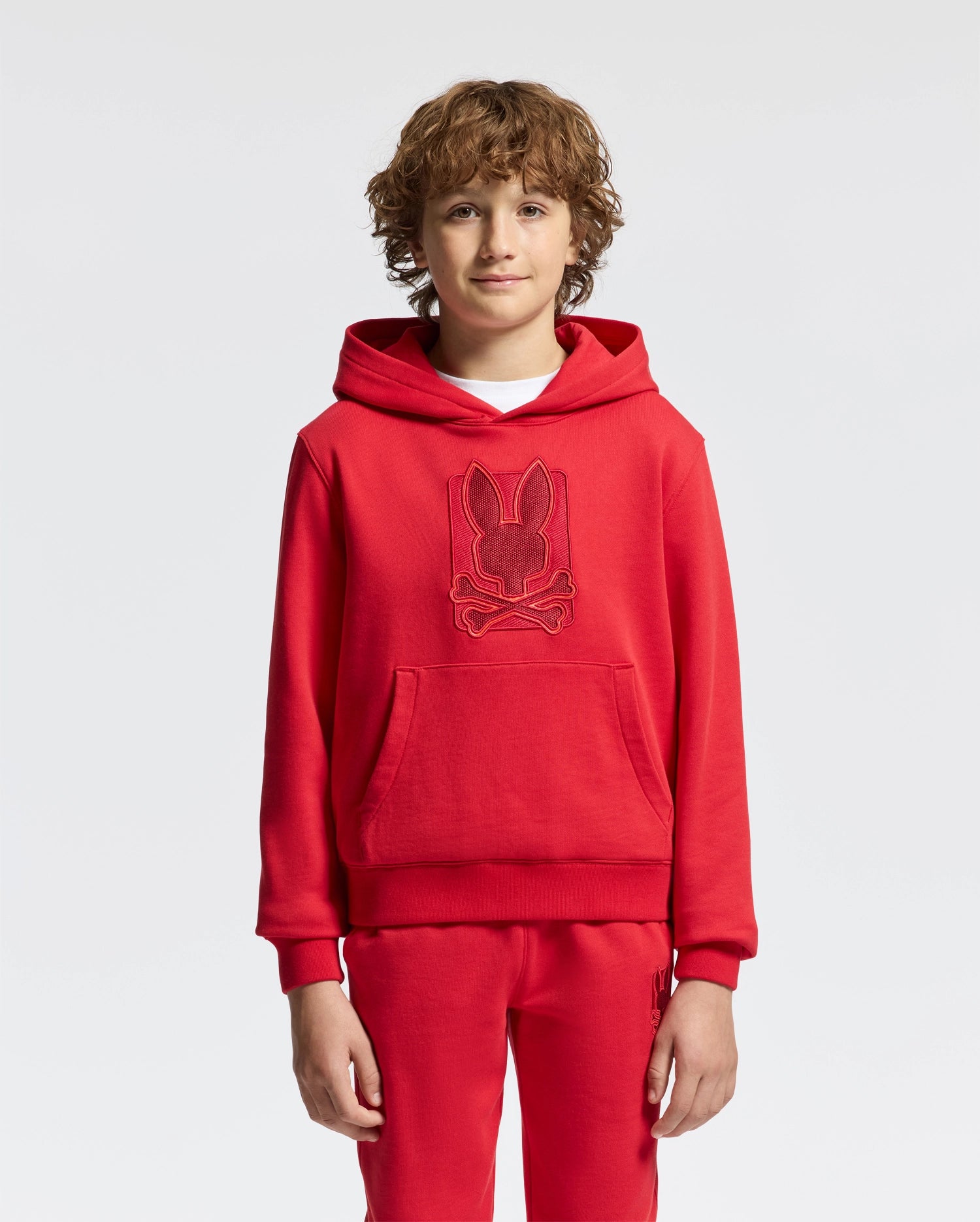 A young person with curly hair is wearing the KIDS PIERCE ESSENTIAL FRENCH TERRY HOODIE from Psycho Bunny, along with matching joggers. The red pullover sweatshirt showcases an embossed design of a bunny with crossed bones beneath it. The plain and well-lit background emphasizes the vibrant kids' cotton ensemble.