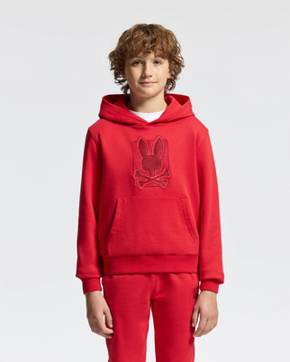 A young person with curly hair is wearing the KIDS PIERCE ESSENTIAL FRENCH TERRY HOODIE from Psycho Bunny, along with matching joggers. The red pullover sweatshirt showcases an embossed design of a bunny with crossed bones beneath it. The plain and well-lit background emphasizes the vibrant kids' cotton ensemble.