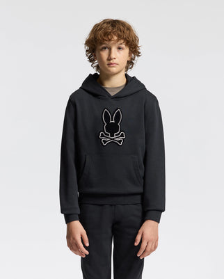 A young person with curly hair models the Psycho Bunny KIDS ALEXANDER HOODIE (B0H154E200), a black hoodie featuring a skull and bunny ears design. This versatile pullover offers a comfortable relaxed fit, standing out against the plain white backdrop.