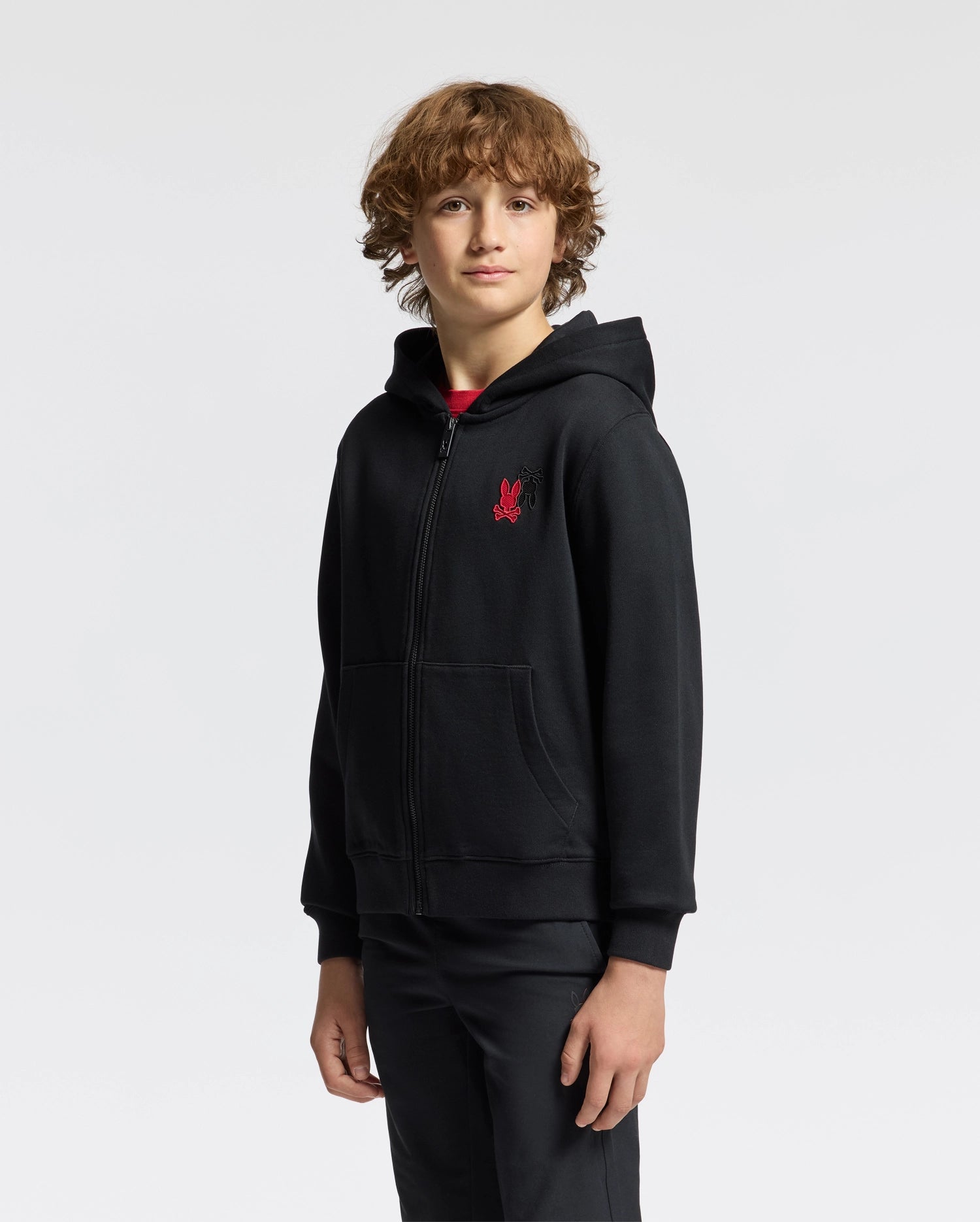 A young person with curly hair stands against a plain background, wearing a Psycho Bunny Kids' Pierce Essential French Terry Zip Hoodie in black, featuring a red logo on the chest, along with black pants. They have a neutral expression and are facing slightly to the side.