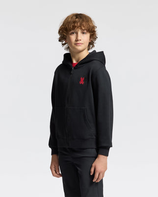 A young person with curly hair stands against a plain background, wearing a Psycho Bunny Kids' Pierce Essential French Terry Zip Hoodie in black, featuring a red logo on the chest, along with black pants. They have a neutral expression and are facing slightly to the side.