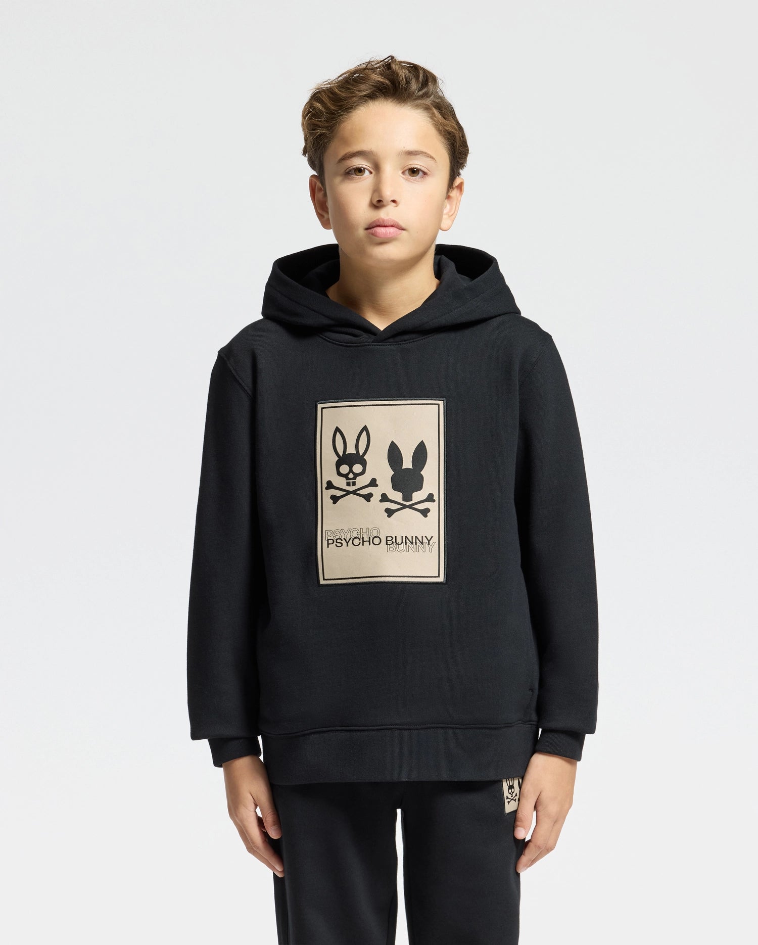 A young boy with light brown hair wears a Psycho Bunny KIDS MONROE TWILL PATCH FLEECE HOODIE featuring a large rectangular logo appliqué with two bunnies. He stands against a plain, light gray background.