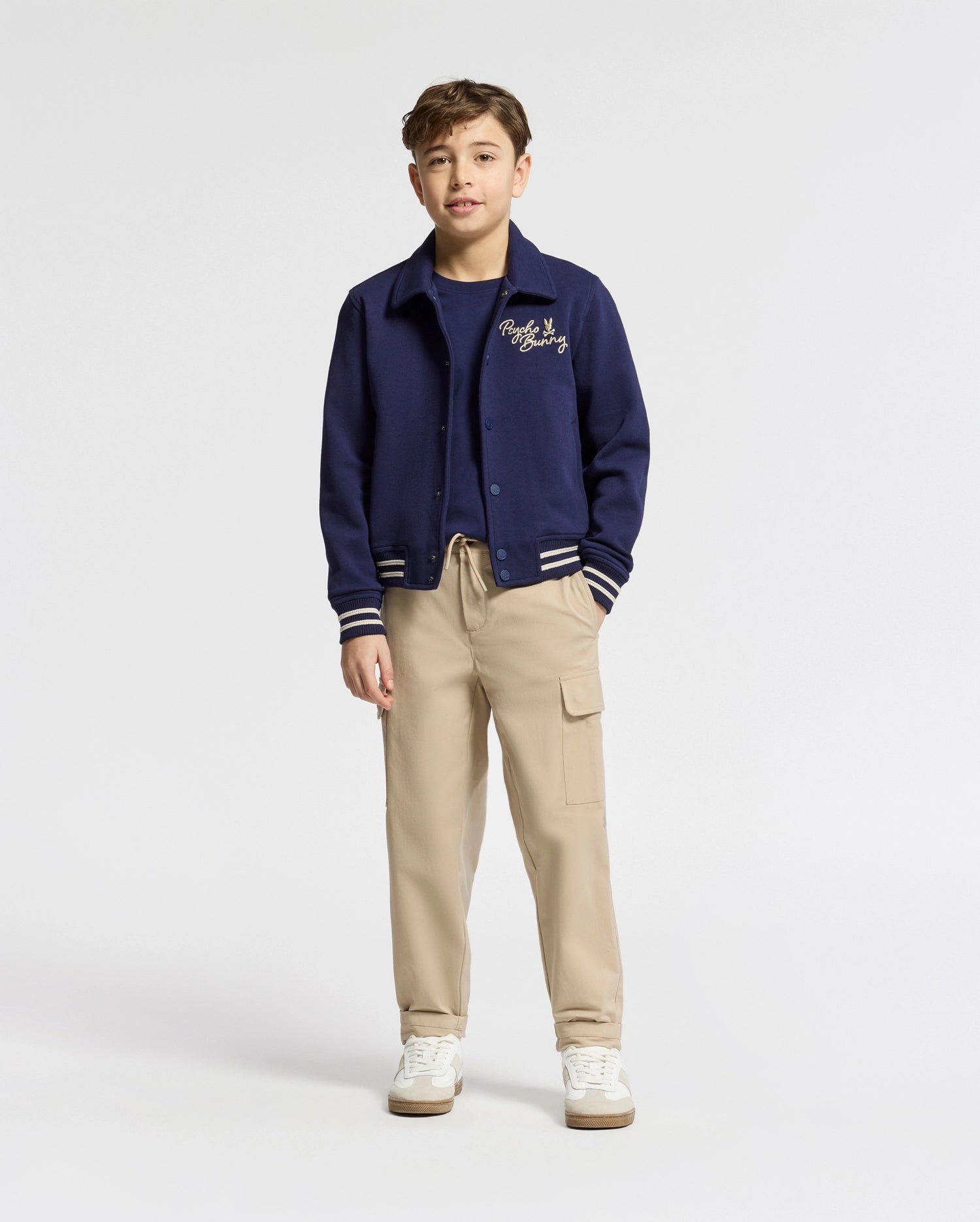 A young person stands confidently, wearing the Psycho Bunny Kids Desert Coach Bomber Jacket - B0J428F200 in Evening Blue with white trim over a navy top and beige cargo pants. Their short hair and white sneakers complete the look against a plain white background.