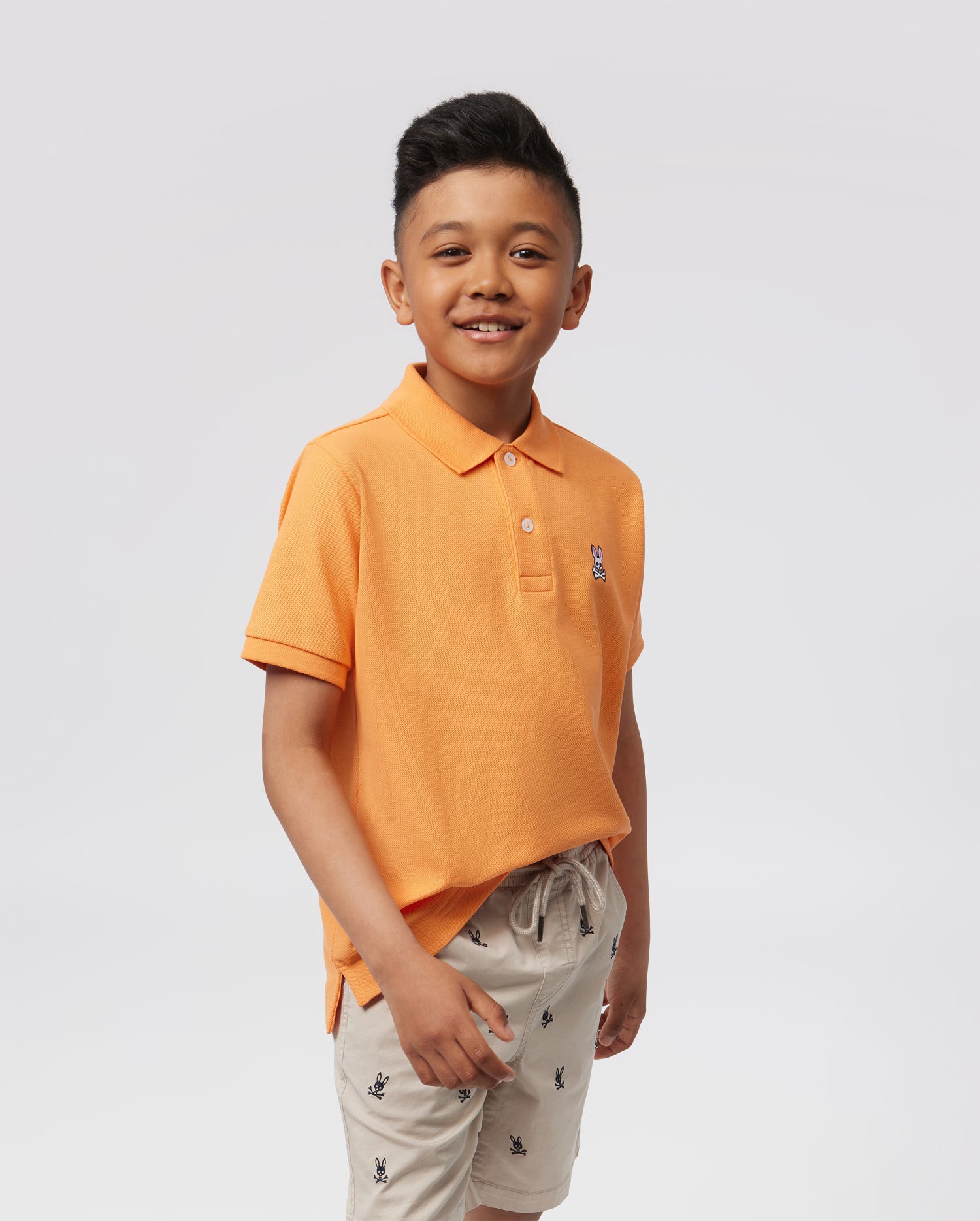 A smiling young boy models the Psycho Bunny KIDS CLASSIC PIQUE POLO - B0K001Z1PC, an orange polo shirt paired with beige shorts featuring small embroidered designs, set against a light background.