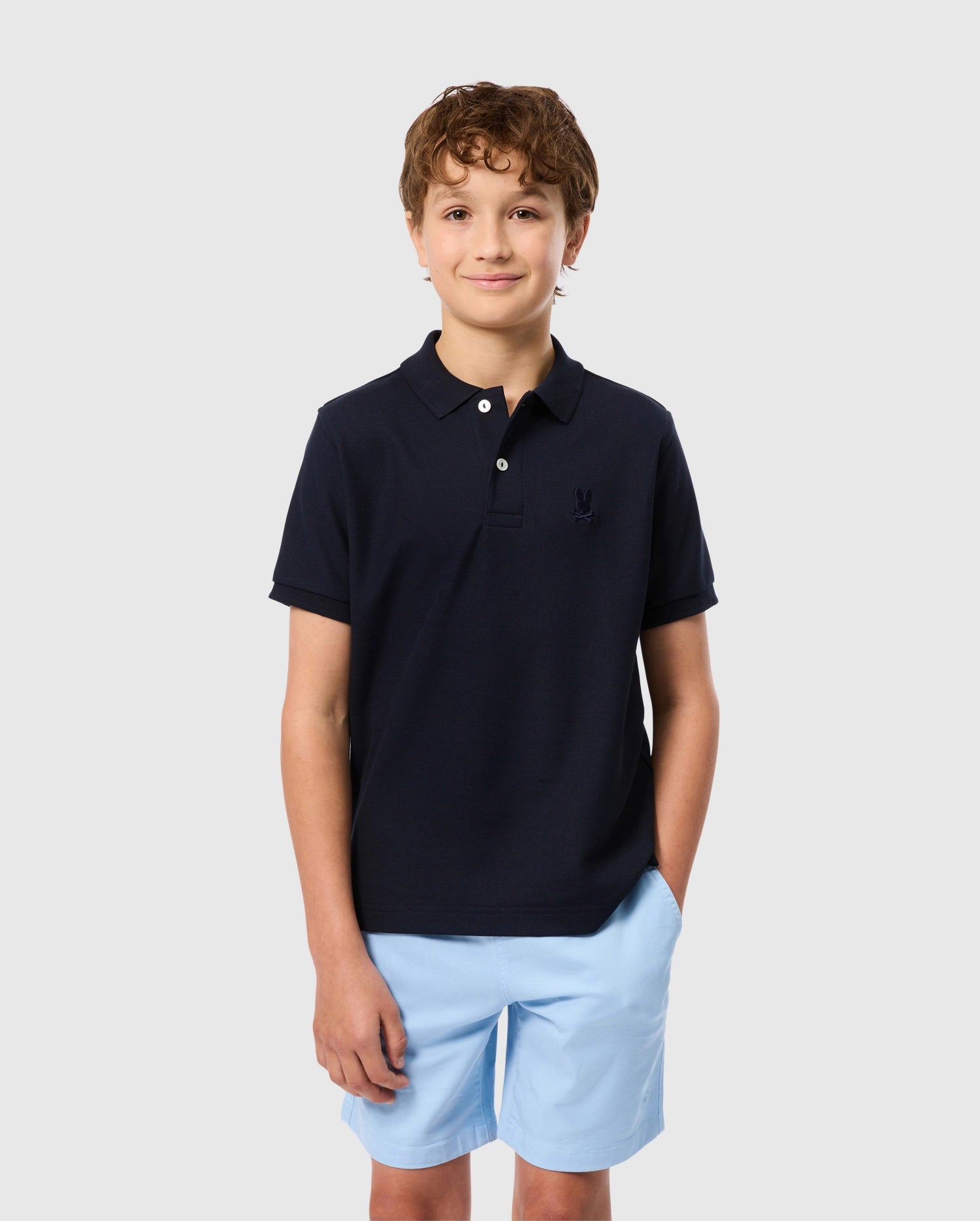 A young boy with curly brown hair stands with his hands in the pockets of his light blue shorts, wearing a Kids Classic Pique Polo Shirt from Psycho Bunny, featuring an embroidered chest Bunny logo. He is smiling slightly and standing against a plain light gray background.