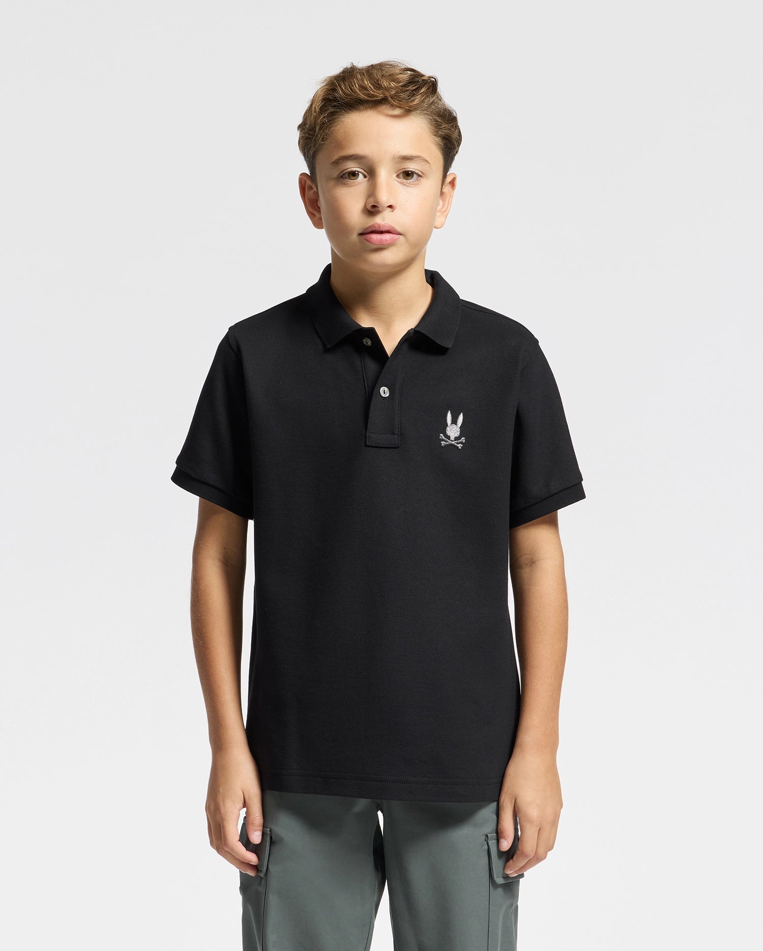 A young boy stands against a plain background, giving off a fashion-forward vibe in his black Psycho Bunny Kids Alexander Pique Polo featuring a subtle white emblem on the chest, paired with gray pants. His short hair complements his neutral expression.