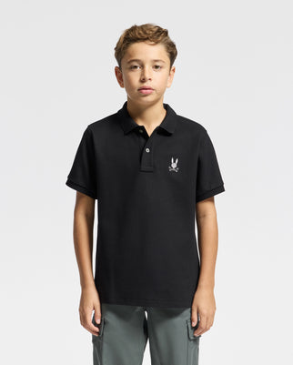 A young boy stands against a plain background, giving off a fashion-forward vibe in his black Psycho Bunny Kids Alexander Pique Polo featuring a subtle white emblem on the chest, paired with gray pants. His short hair complements his neutral expression.