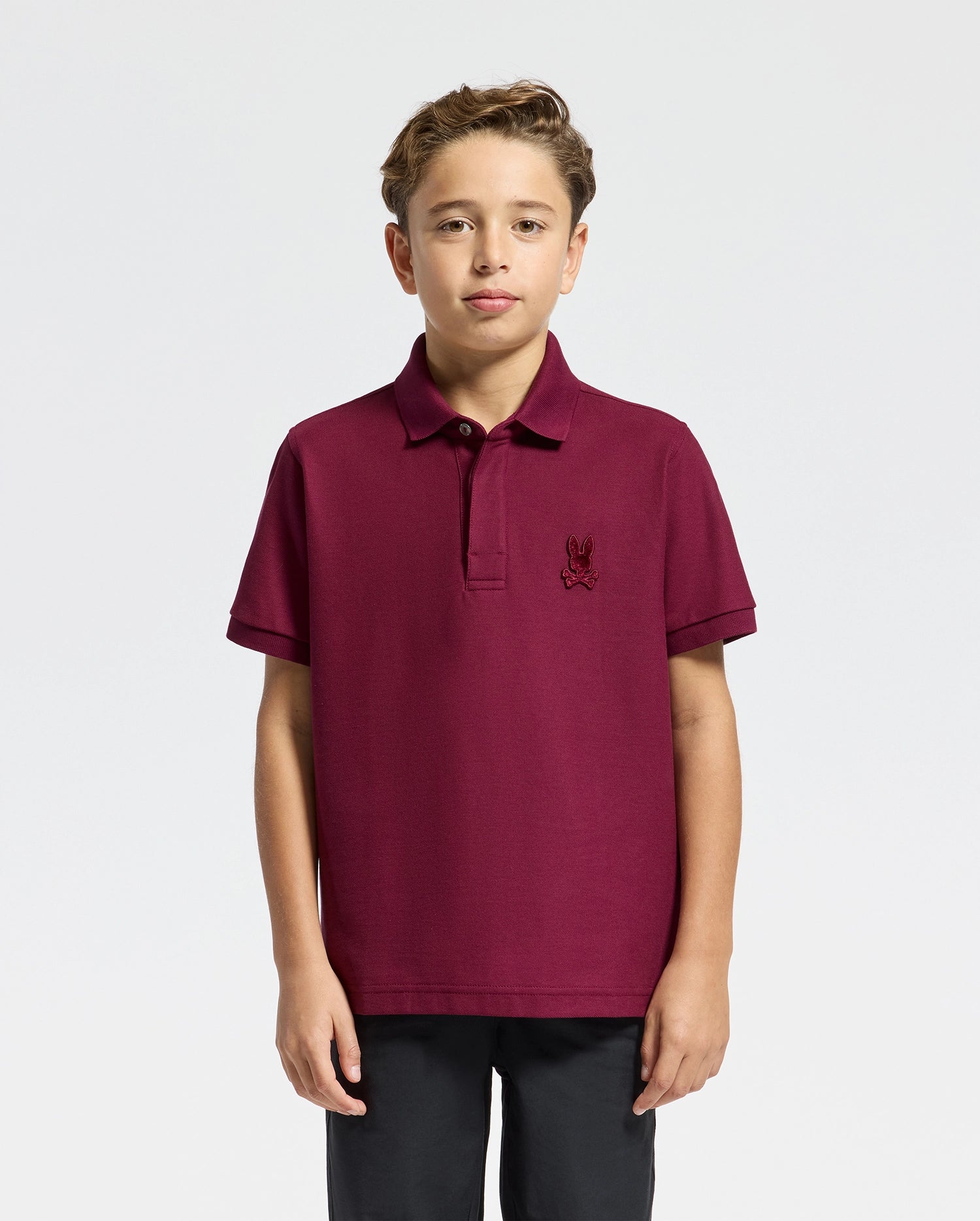 A young boy stands wearing a maroon Psycho Bunny Kids Brandson Pique Polo with an embroidered logo on the chest, paired with black pants. The background is plain and light-colored.