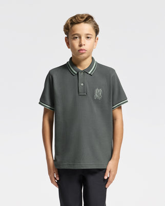 A young boy with short blonde hair is wearing a stylish KIDS PIERCE PIQUE POLO from Psycho Bunny, made from soft Pima cotton. The dark gray shirt showcases green striped edges and an embroidered chest Bunny logo. He stands against a plain white background, looking directly at the camera.