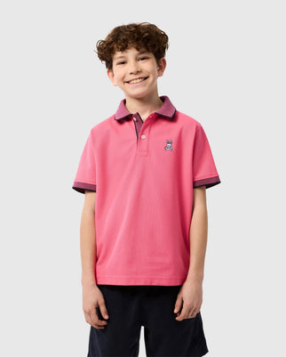 A smiling young boy with curly hair stands against a plain background. He is wearing a Psycho Bunny KIDS SOUTHPORT PIQUE POLO SHIRT - B0K263B200 made from Pima cotton piqué, featuring a small embroidered bunny logo on the left chest and dark shorts. The collar and sleeve edges of the shirt contrast beautifully with the pink.