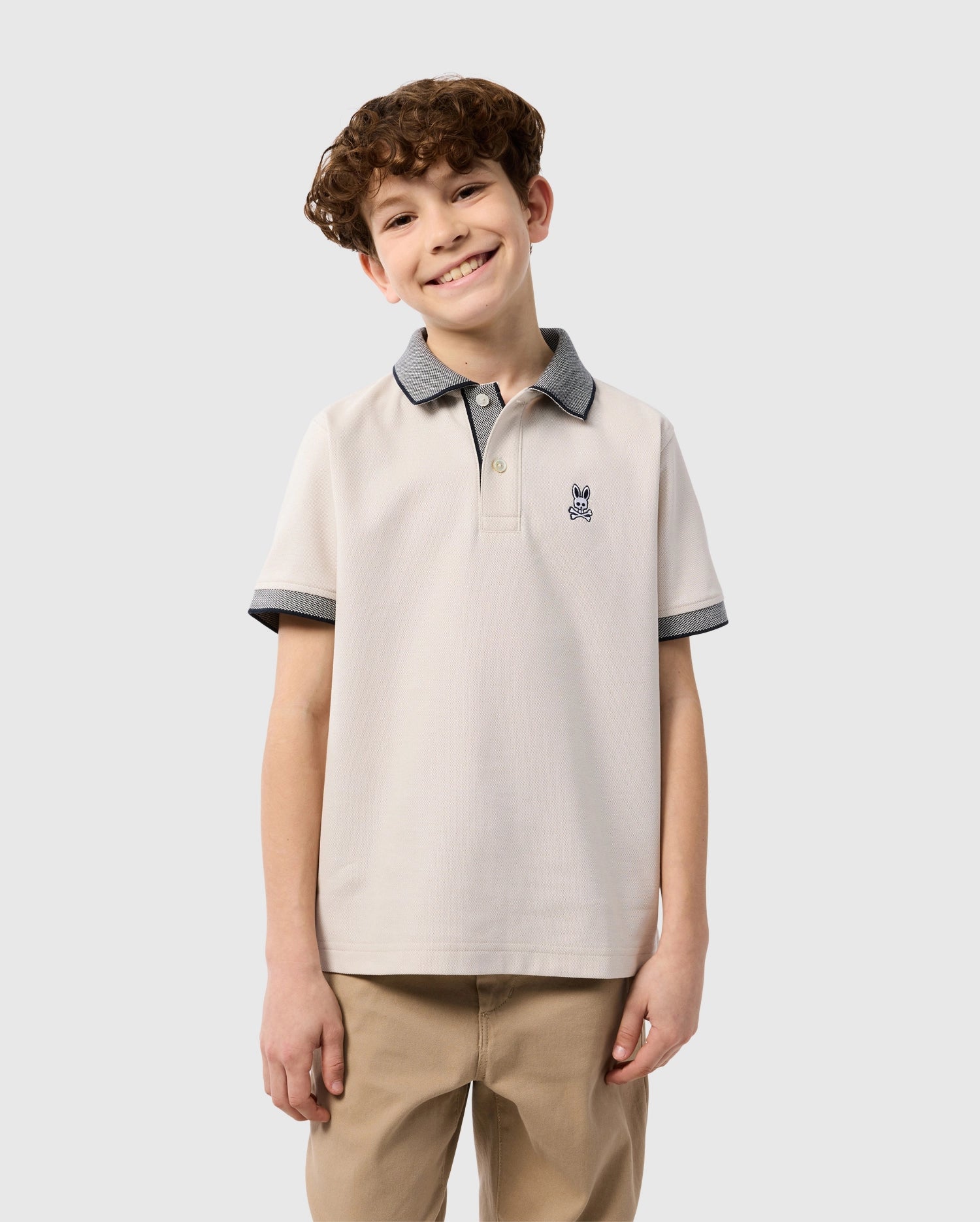 A smiling young boy with curly brown hair is wearing a beige KIDS SOUTHPORT PIQUE POLO SHIRT - B0K263B200 by Psycho Bunny made of Pima cotton piqué with grey collar and sleeve trim, and khaki pants. He stands against a plain light grey background.