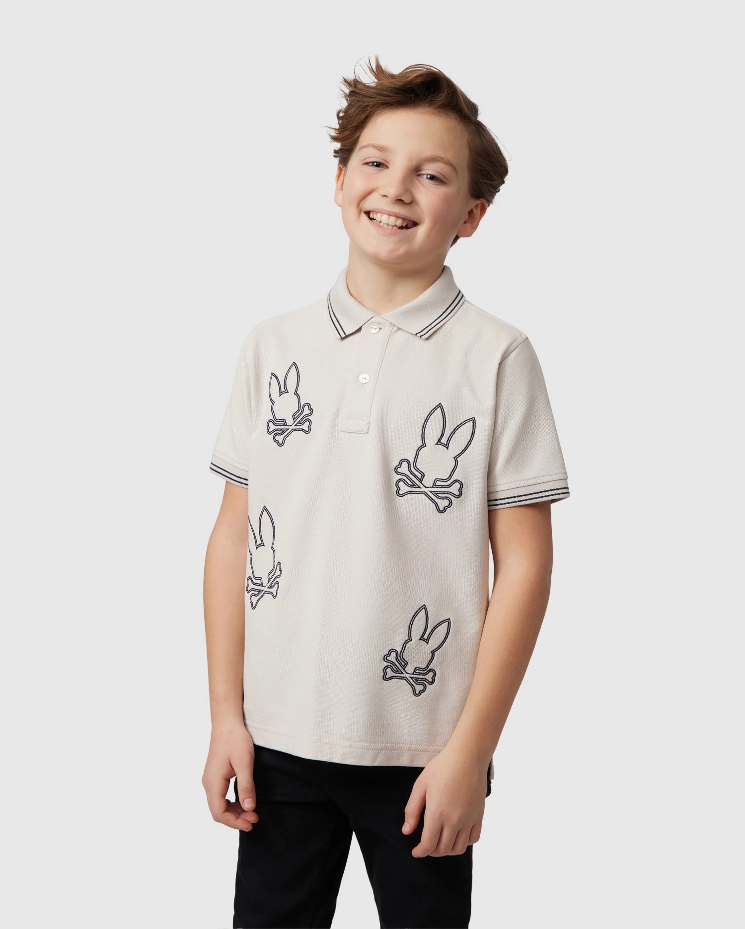 A young boy with light brown hair and a smile is wearing a Psycho Bunny KIDS MILTON PIQUE POLO SHIRT - B0K284B200 adorned with black, stylized bunny skull designs. The beige Pima cotton piqué shirt has a buttoned collar and black trim on the sleeves. He stands against a plain, light gray background.