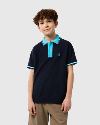 A young boy with curly hair stands smiling at the camera, wearing a navy blue KIDS SALINA PIQUE POLO SHIRT by Psycho Bunny with light blue and white trim and a small logo on the left chest. He is also wearing tan pants.