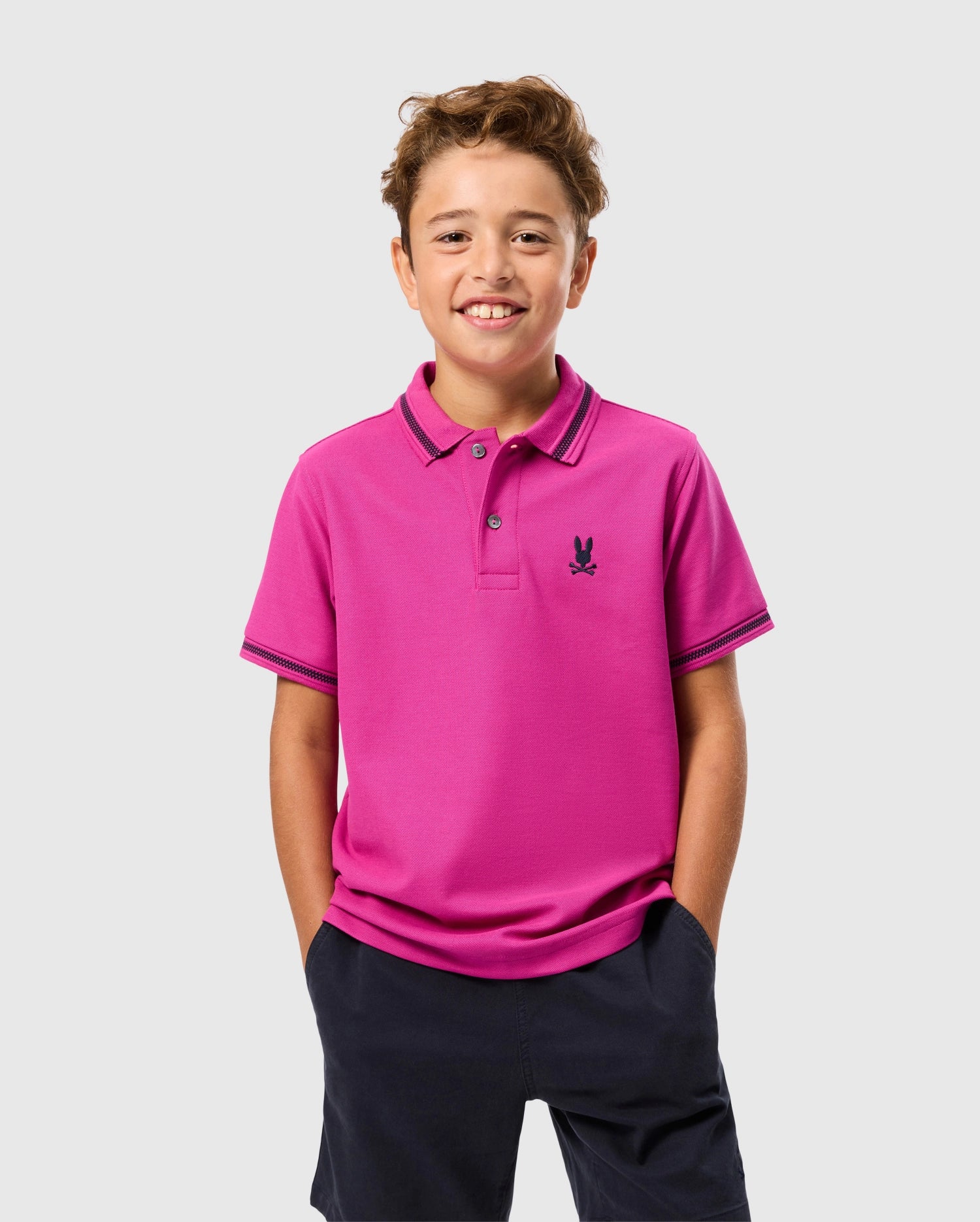 A smiling boy stands with his hands in the pockets of his navy shorts, wearing a bright pink KIDS RIVIERA PIQUE POLO SHIRT by Psycho Bunny. Crafted from Pima cotton piqué, the polo features an embroidered chest Bunny emblem against a plain light gray background.