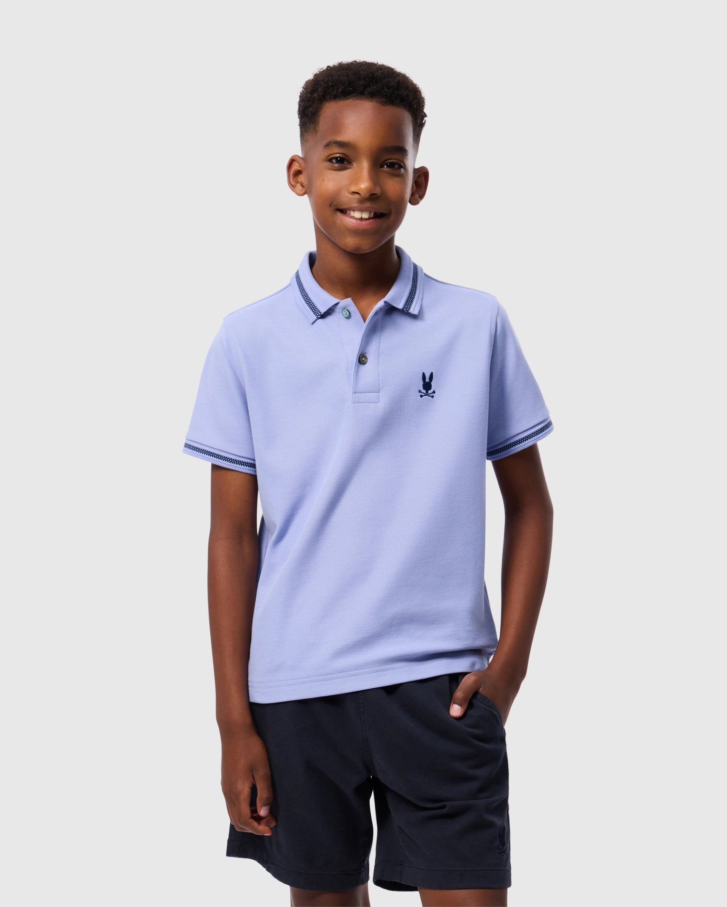 A young boy is smiling while wearing a light blue Psycho Bunny KIDS RIVIERA PIQUE POLO SHIRT (B0K640C200) made of soft Pima cotton piqué, with an embroidered chest Bunny emblem. He pairs it with black shorts, casually resting one hand in his pocket as he stands against a plain gray background.