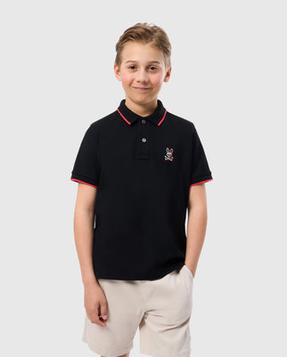 A smiling young boy stands against a plain background, hands in pockets. He is wearing a Psycho Bunny Kids Kayden Pique Polo (B0K675C200) in black with bold contrasting stripes on the collar and sleeves, featuring a small embroidered bunny logo on the left chest, paired with light-colored shorts.