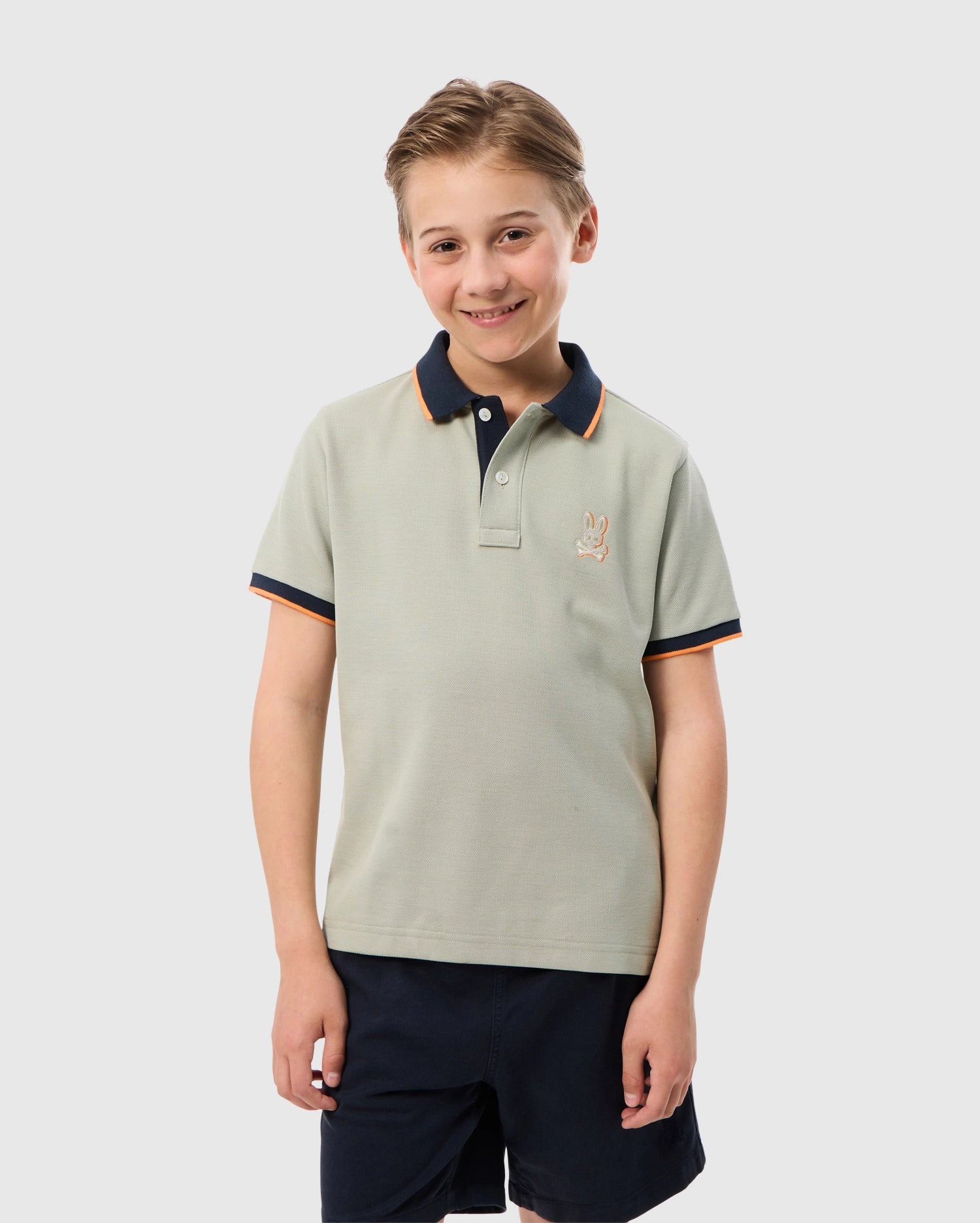 A young boy with short, light brown hair stands smiling. He is wearing a light gray KIDS KAYDEN PIQUE POLO - B0K675C200 by Psycho Bunny, featuring a small bunny on the chest, a navy blue collar, and contrasting stripes as trim. He has paired it with dark shorts against a plain white background.