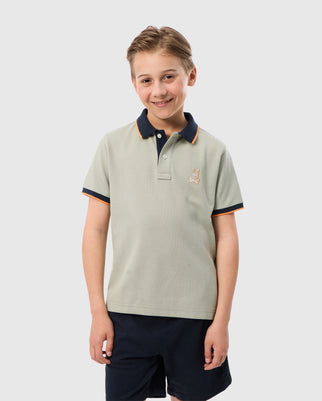 A young boy with short, light brown hair stands smiling. He is wearing a light gray KIDS KAYDEN PIQUE POLO - B0K675C200 by Psycho Bunny, featuring a small bunny on the chest, a navy blue collar, and contrasting stripes as trim. He has paired it with dark shorts against a plain white background.