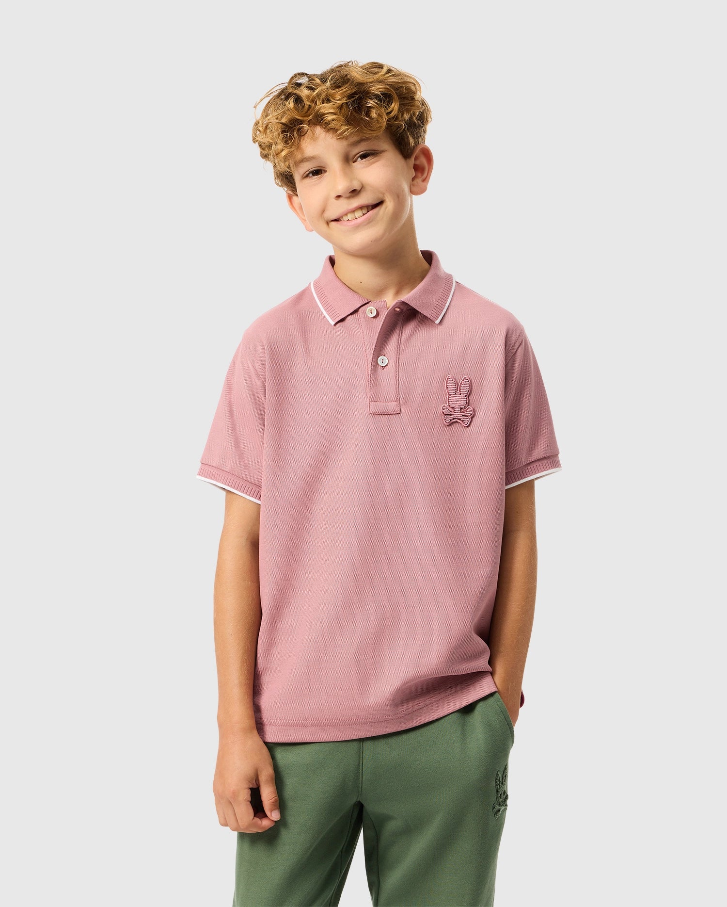 A smiling young person with curly hair is wearing a pink KIDS KINGSTON PIQUE POLO SHIRT from Psycho Bunny, crafted from Pima cotton and featuring an embroidered Bunny patch. They pair it with green pants, standing confidently with one hand in their pocket against a plain background.