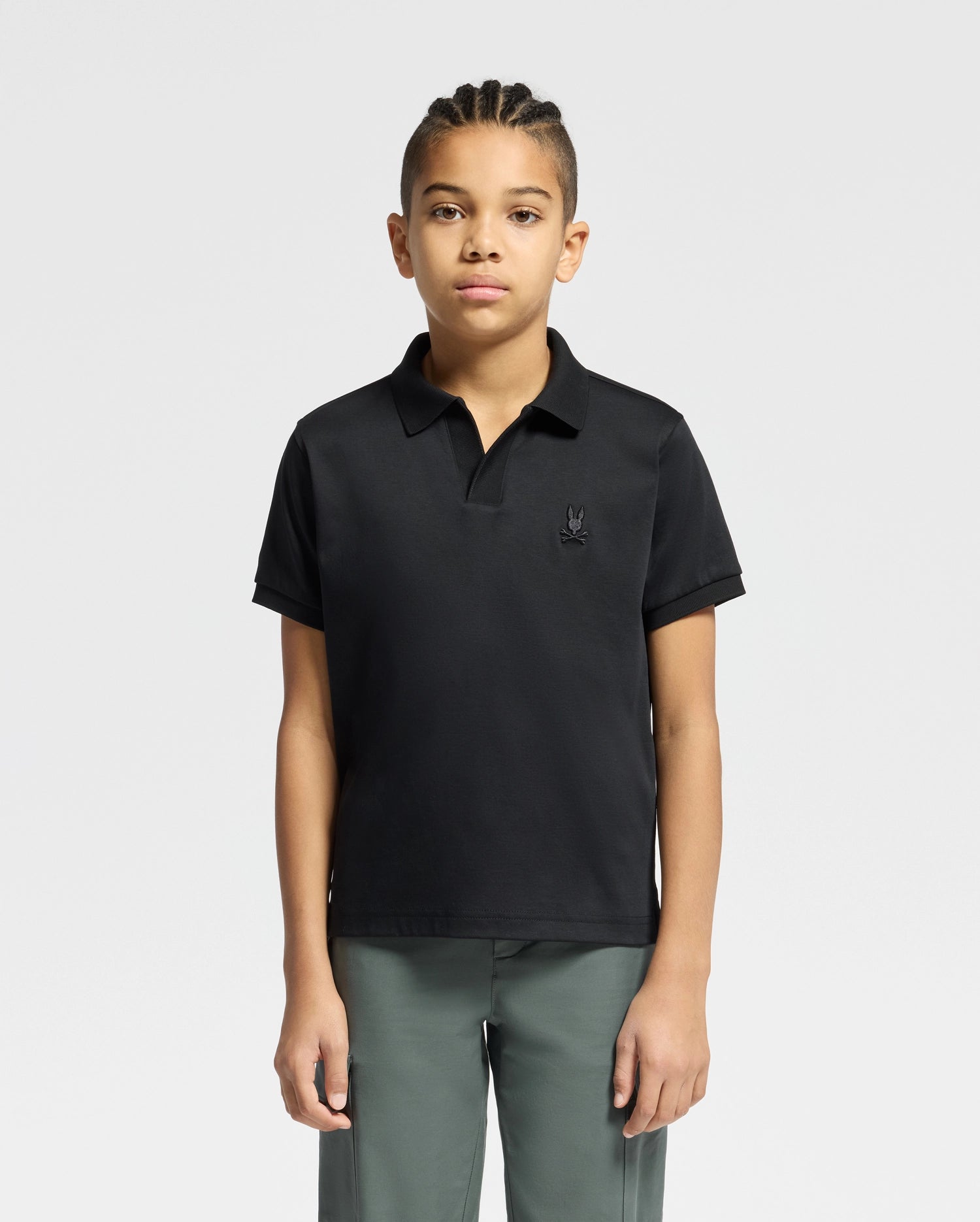 A young person with braided hair is wearing a black Psycho Bunny KIDS BROADWAY PIMA INTERLOCK JOHNNY COLLAR POLO and gray pants, standing against a plain white background.