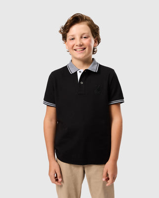 A young boy with curly hair, wearing a Psycho Bunny KIDS MALTON PIQUE POLO SHIRT - B0K950D200 made from Pima cotton piqué with a grey collar and tan pants, smiles and stands against a plain grey background.