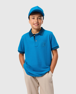 A boy smiles while wearing a blue Psycho Bunny Kids Malton Pique Polo Shirt (B0K950D200) made of Pima cotton piqué, featuring a small embroidered logo on the chest and mother-of-pearl buttons. He has a matching blue baseball cap and his hands tucked in the pockets of his beige pants against a plain light gray background.