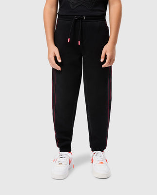 A child is wearing an elastic and comfortable black t-shirt paired with the KIDS TRAVIS TRACK PANT - B0P143D200 by Psycho Bunny, featuring pink contrast chainstitch stripes down the sides. They have on white sneakers with pink accents against a plain, light-colored background.